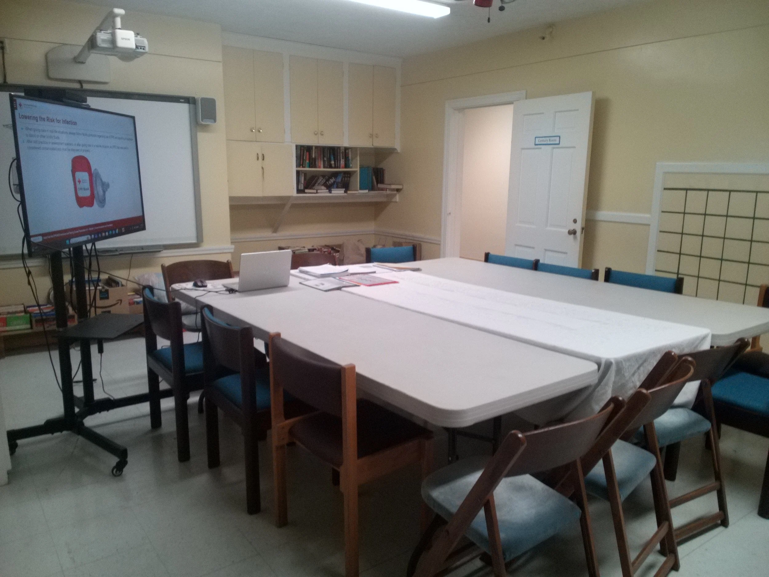 Empty conference room with a large table covered in a white cloth, surrounded by mixed wooden and padded chairs. A large monitor on a stand displays a presentation on health and safety. There are shelves with books and a white door labeled 'Community