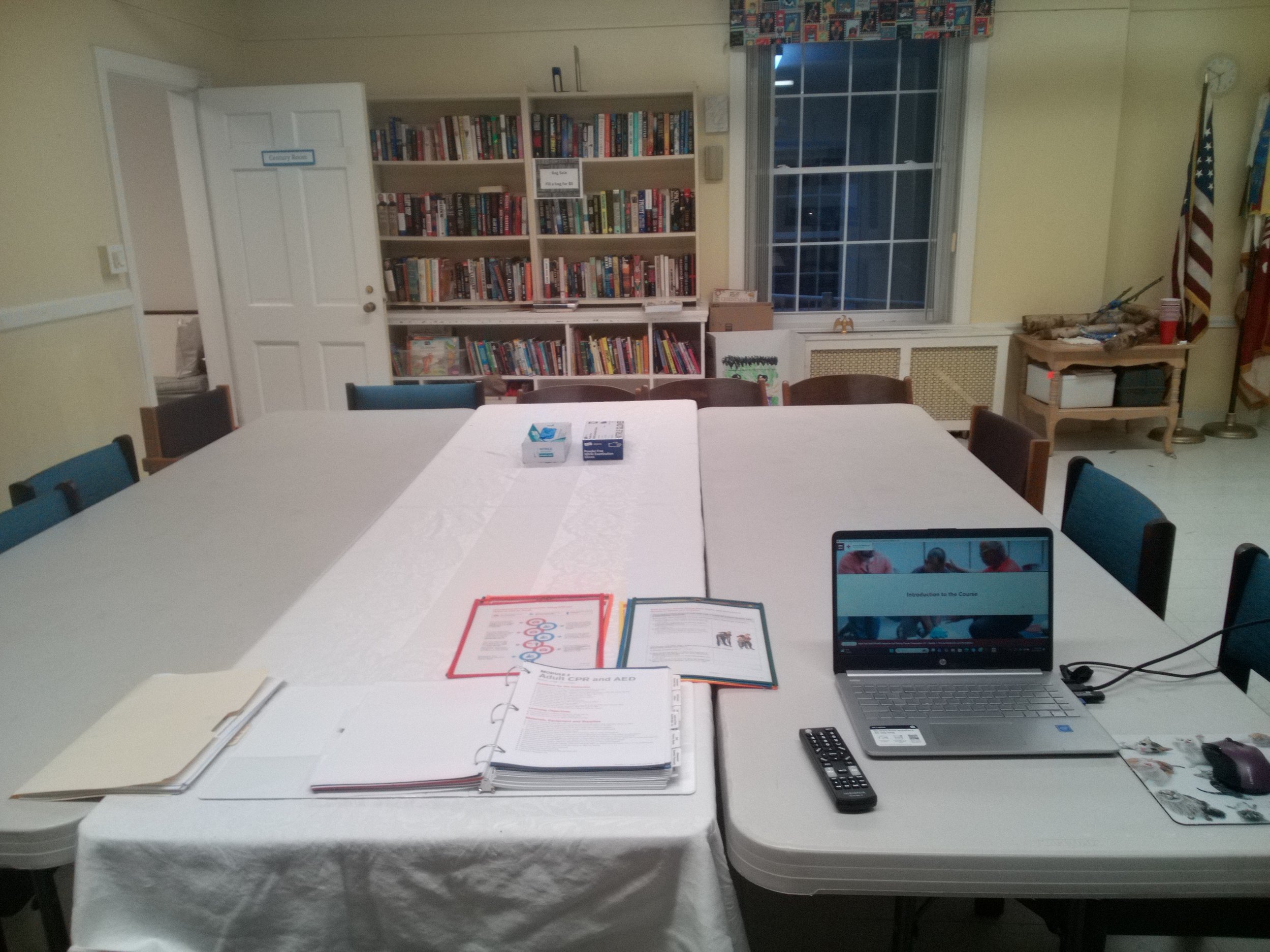 A conference room with a large table covered with a white tablecloth, electronic equipment, papers, and binders, surrounded by chairs, with a bookshelf filled with books and a window in the background.