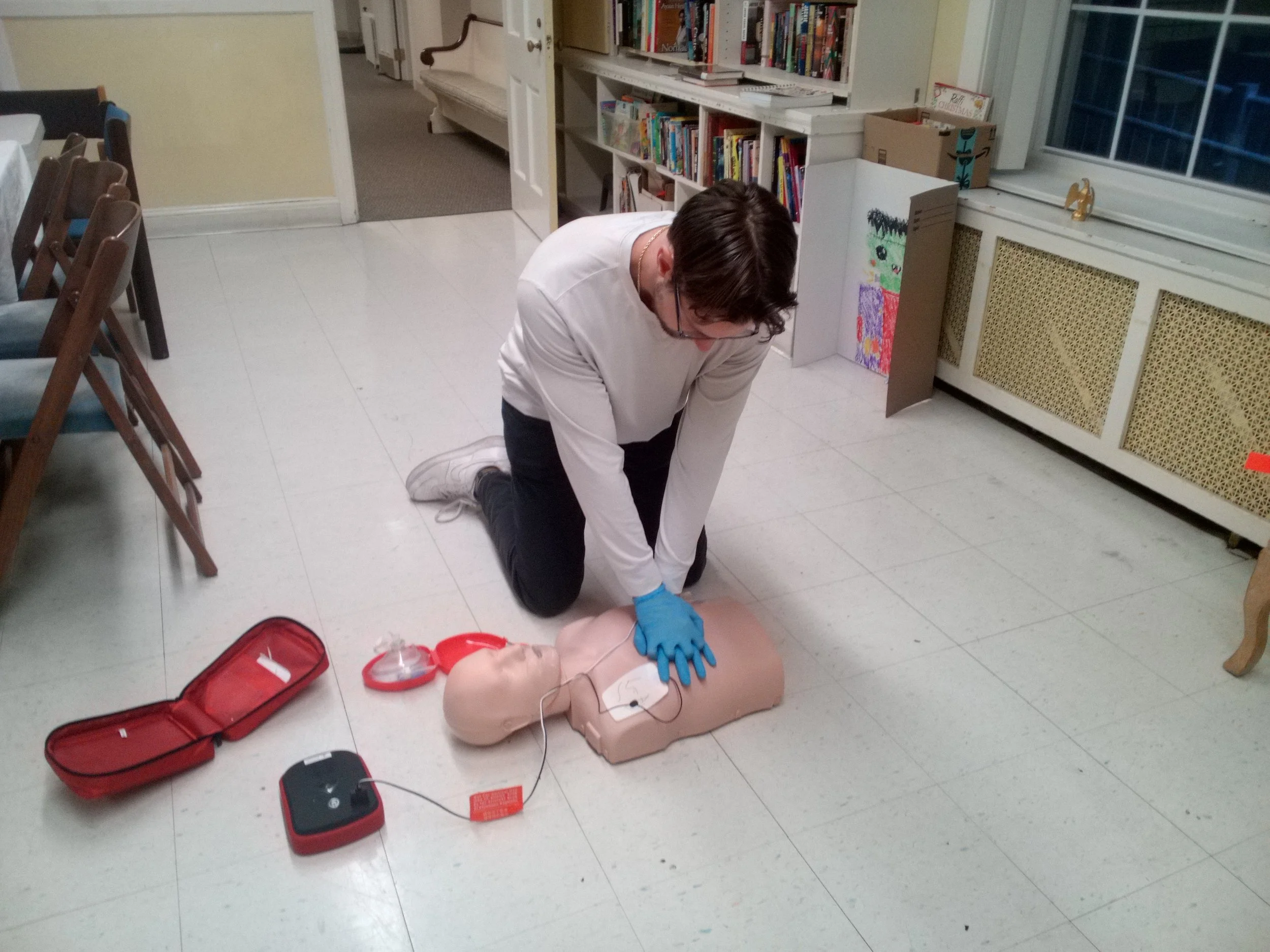 Person practicing CPR on a training mannequin in a room with bookshelves and chairs.