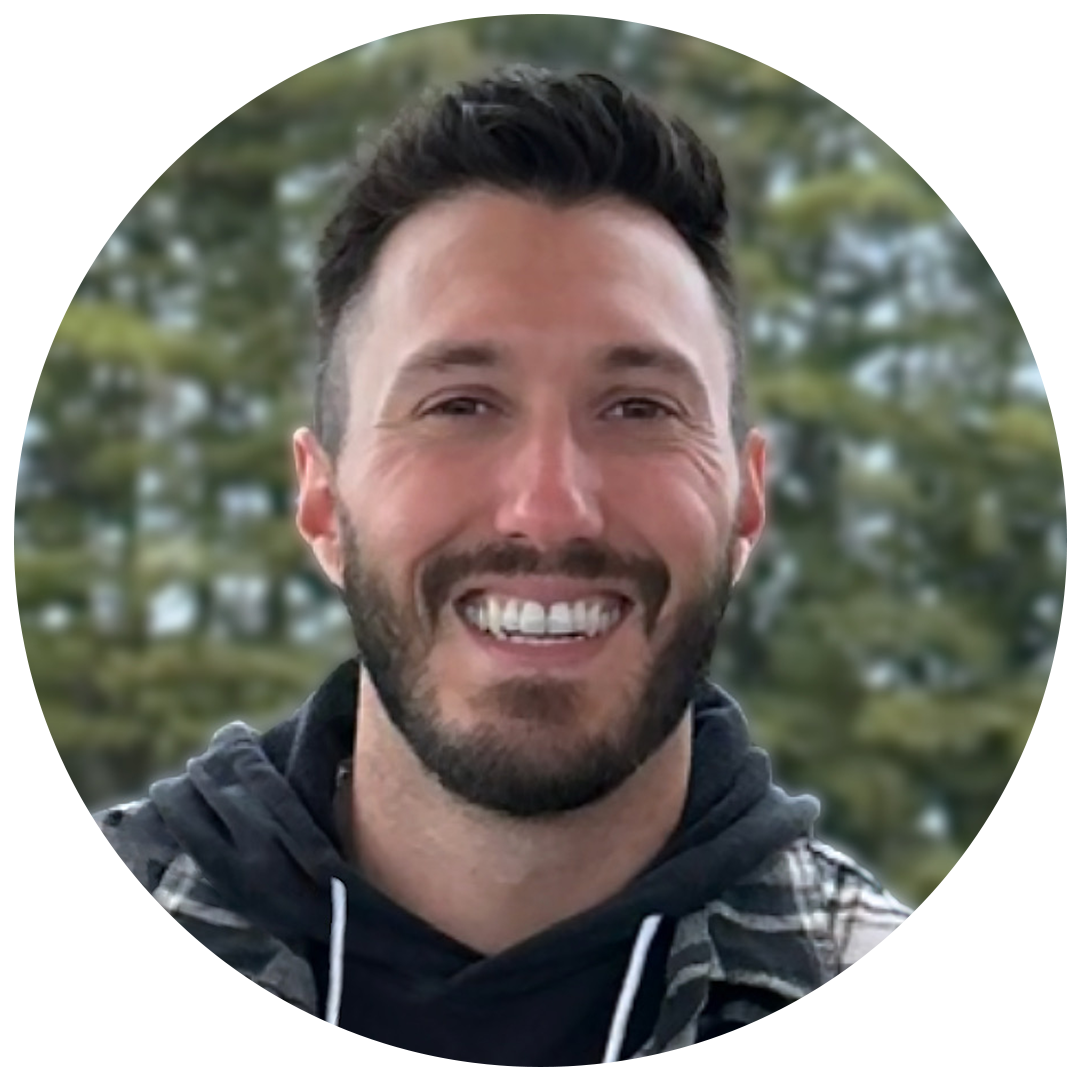 Smiling man with dark hair and beard, wearing a black hoodie and a jacket, outdoors with blurred trees in the background.