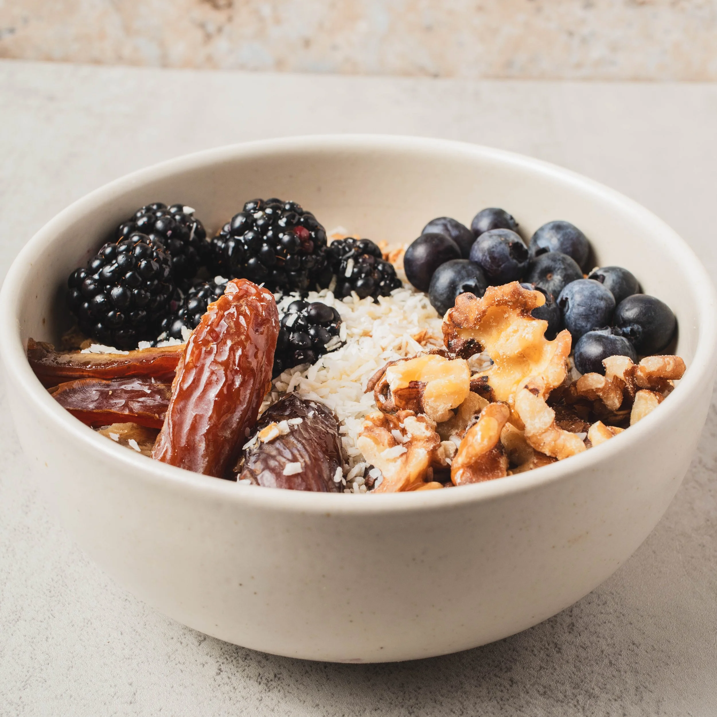 White bowl filled with blackberries, blueberries, chopped coconut, dates, and walnuts on a light gray surface.