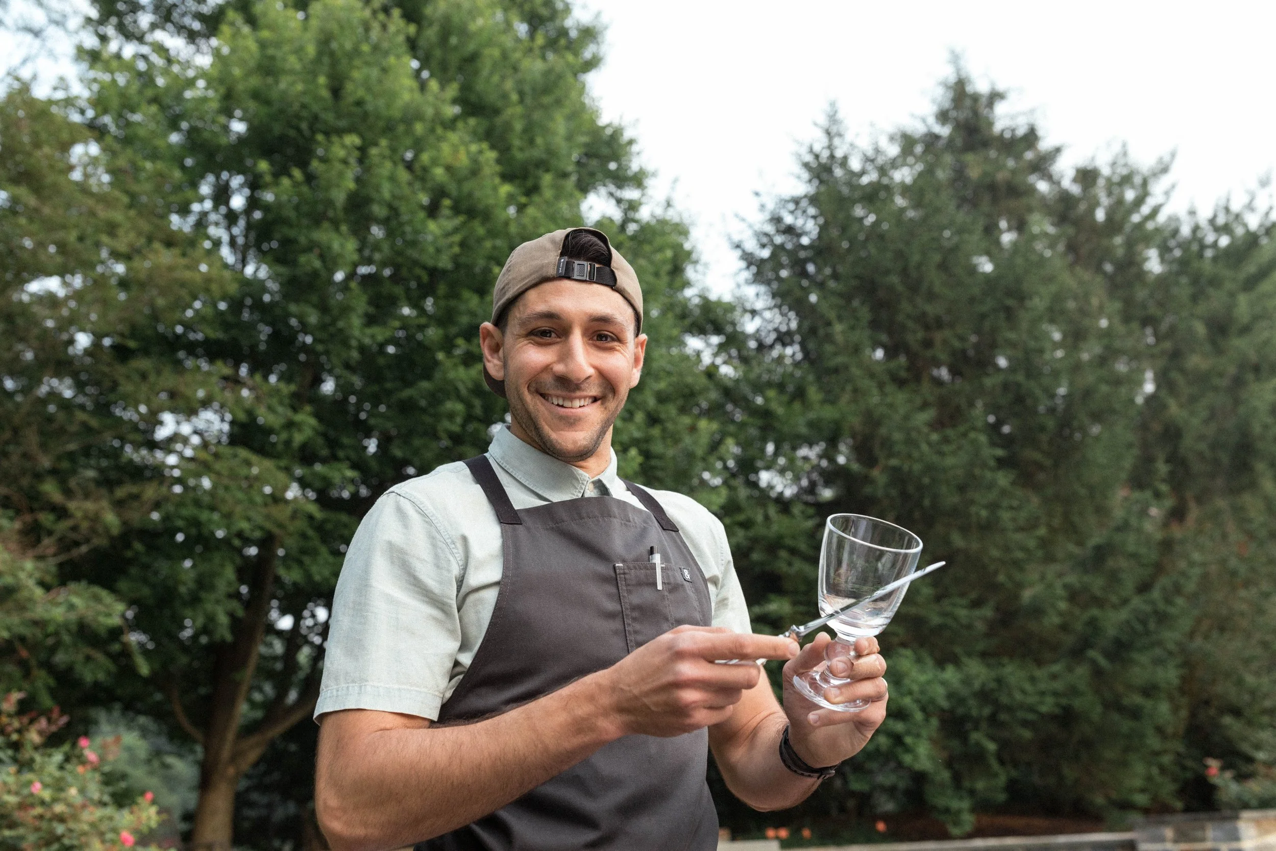 Smiling man wearing a bartender apron and baseball cap holding a wine glass and a fork outdoors with trees in the background.