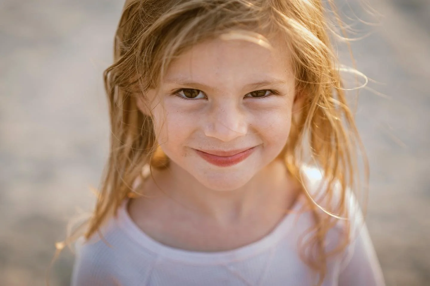 Close-up of a young girl with red hair smiling at camera, outdoors in sunlight.