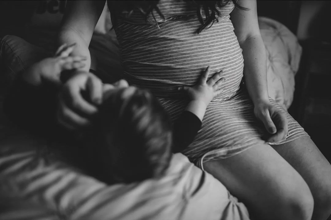 A maternity photo of a mother at home with her son. Her son is touching his mom's belly