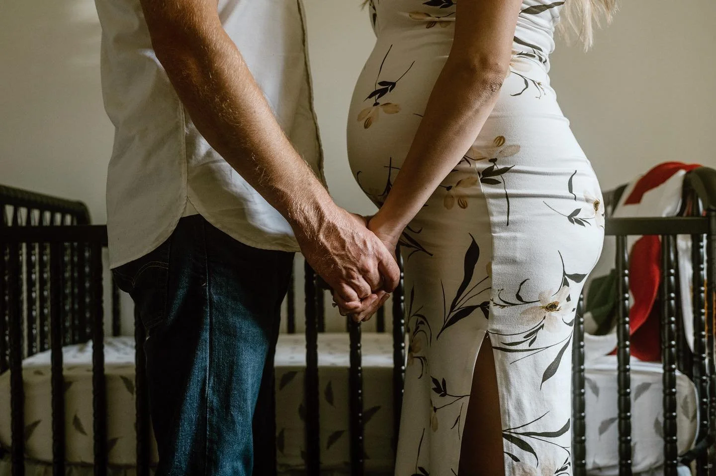 A maternity style photo of a mom and dad in their nursery holding hands. The mom has a pregnant belly