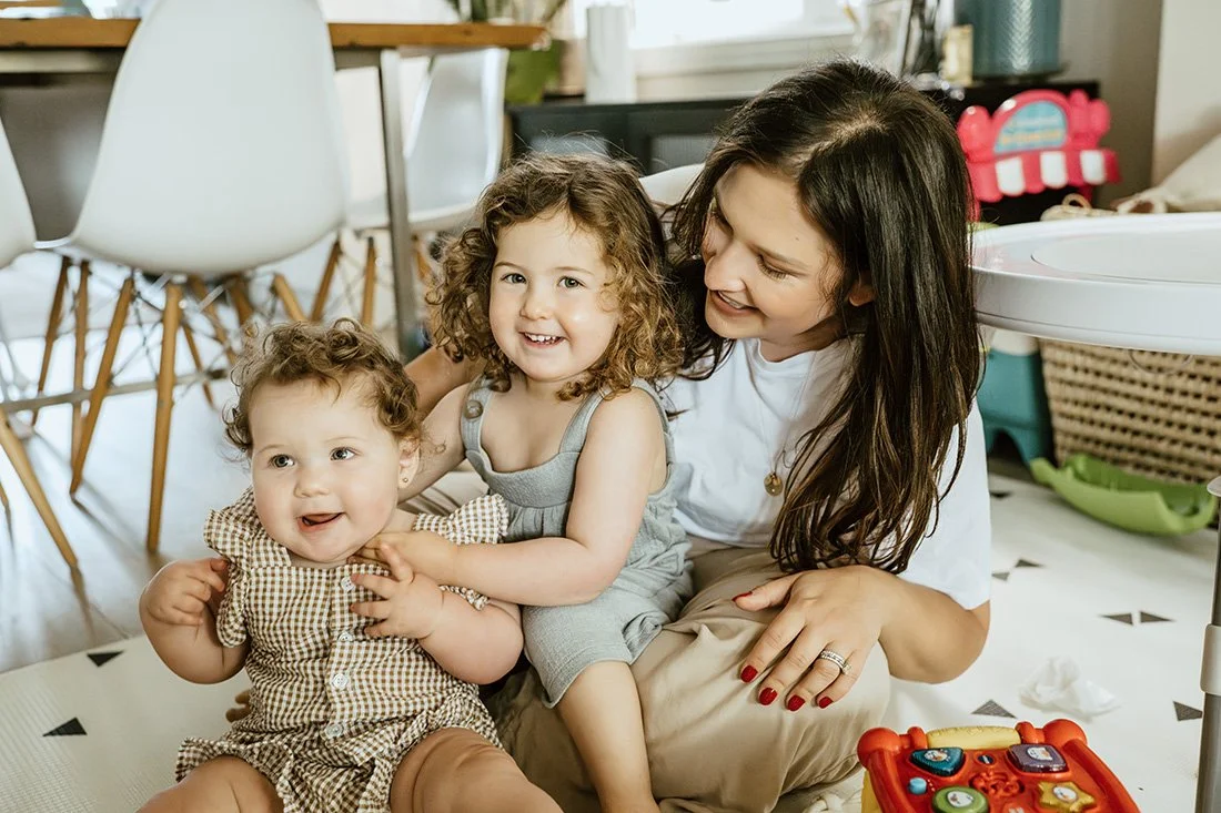 Mom playing with daughters on the floor of a kitchen, smiling and enjoying a moment together.