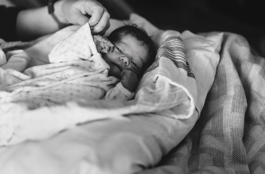 A newborn baby lying in a Mount Sinai hospital bassinet, covered with blankets, being gently cared for by mom's hand