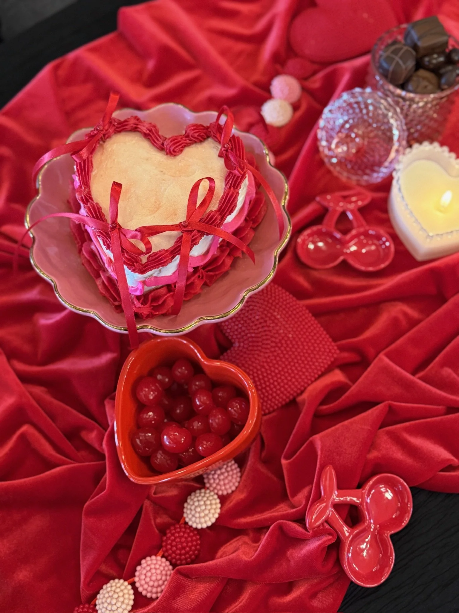 Valentine's Day themed table setting with a heart-shaped cake topped with pink and red icing ribbons, surrounded by red heart-shaped bowls filled with cherries, candles, chocolates, and pink and white decorative candies on red velvet fabric.
