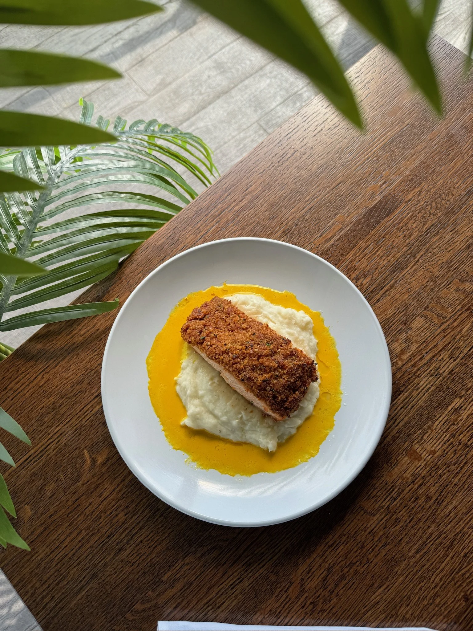 A plate with mashed potatoes topped with a breaded and fried piece of fish, surrounded by a yellow sauce or gravy.