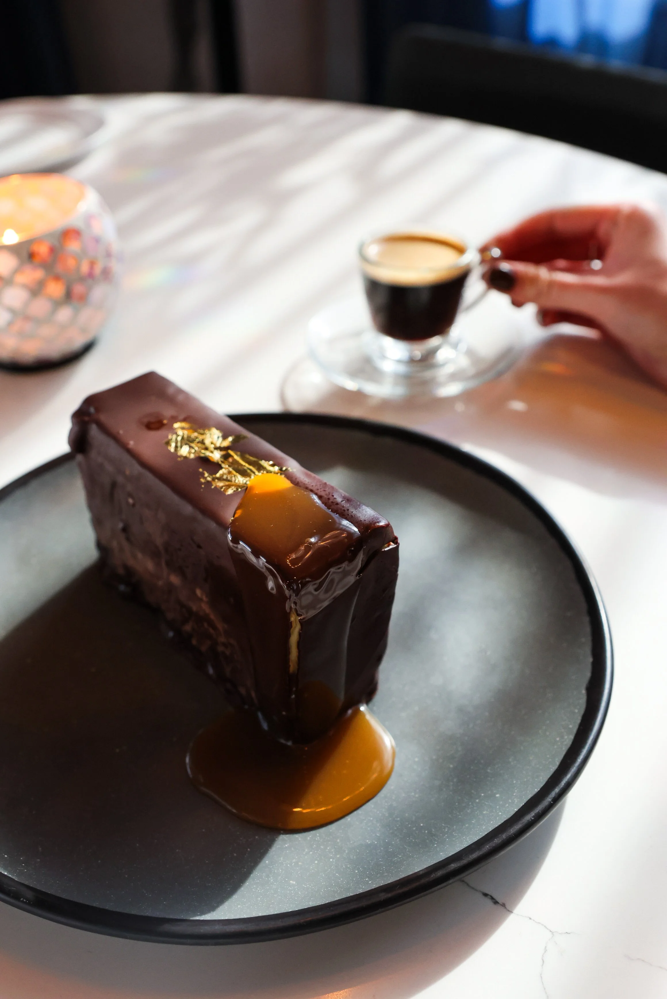 A slice of chocolate cake with caramel sauce on a black plate, a small cup of espresso, and a hand holding a small cup of coffee or espresso on a white table.