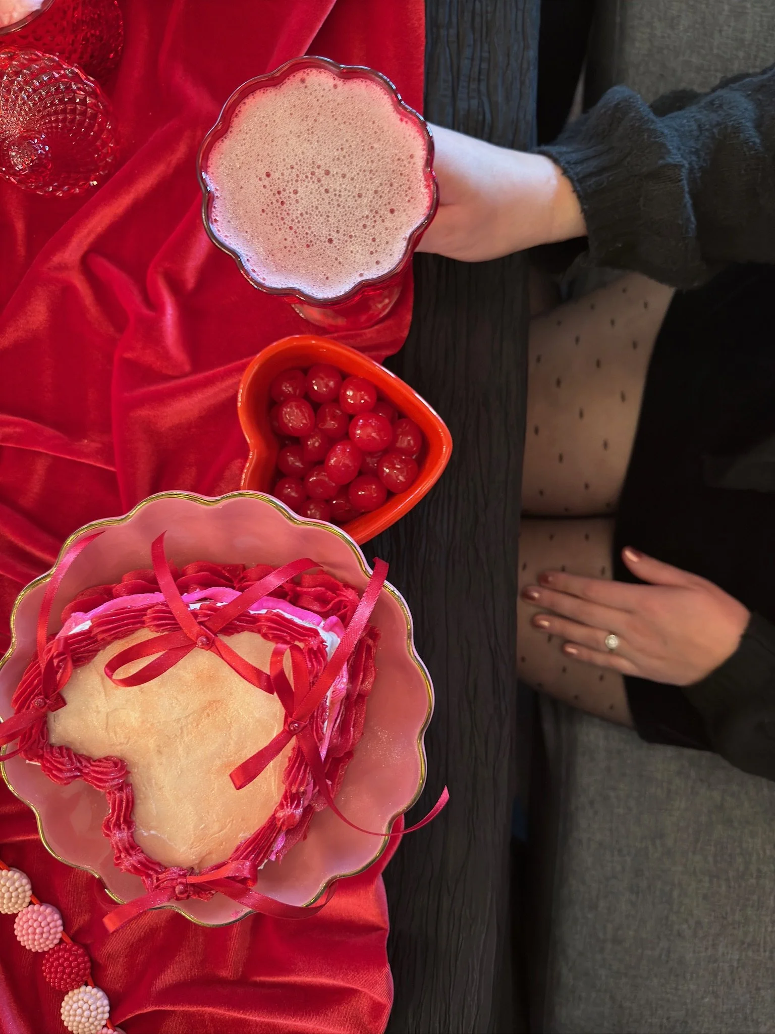 A table with a red satin tablecloth holds a glass of pink frothy drink, a bowl of shiny red cherries, and a heart-shaped cake decorated with pink and red ribbons, with a woman seated nearby wearing black polka dot tights and a black sweater.