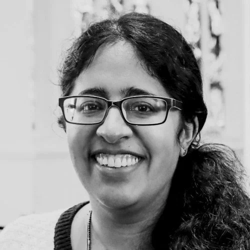 A smiling South Asian woman with glasses and curly hair, wearing a sweater, indoors.