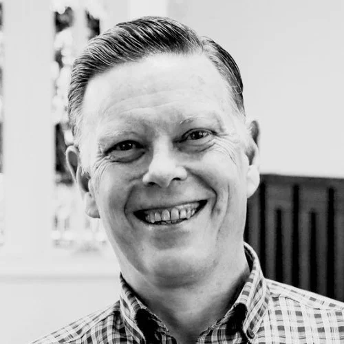 A smiling man with neatly combed hair and a checkered shirt, in black and white.