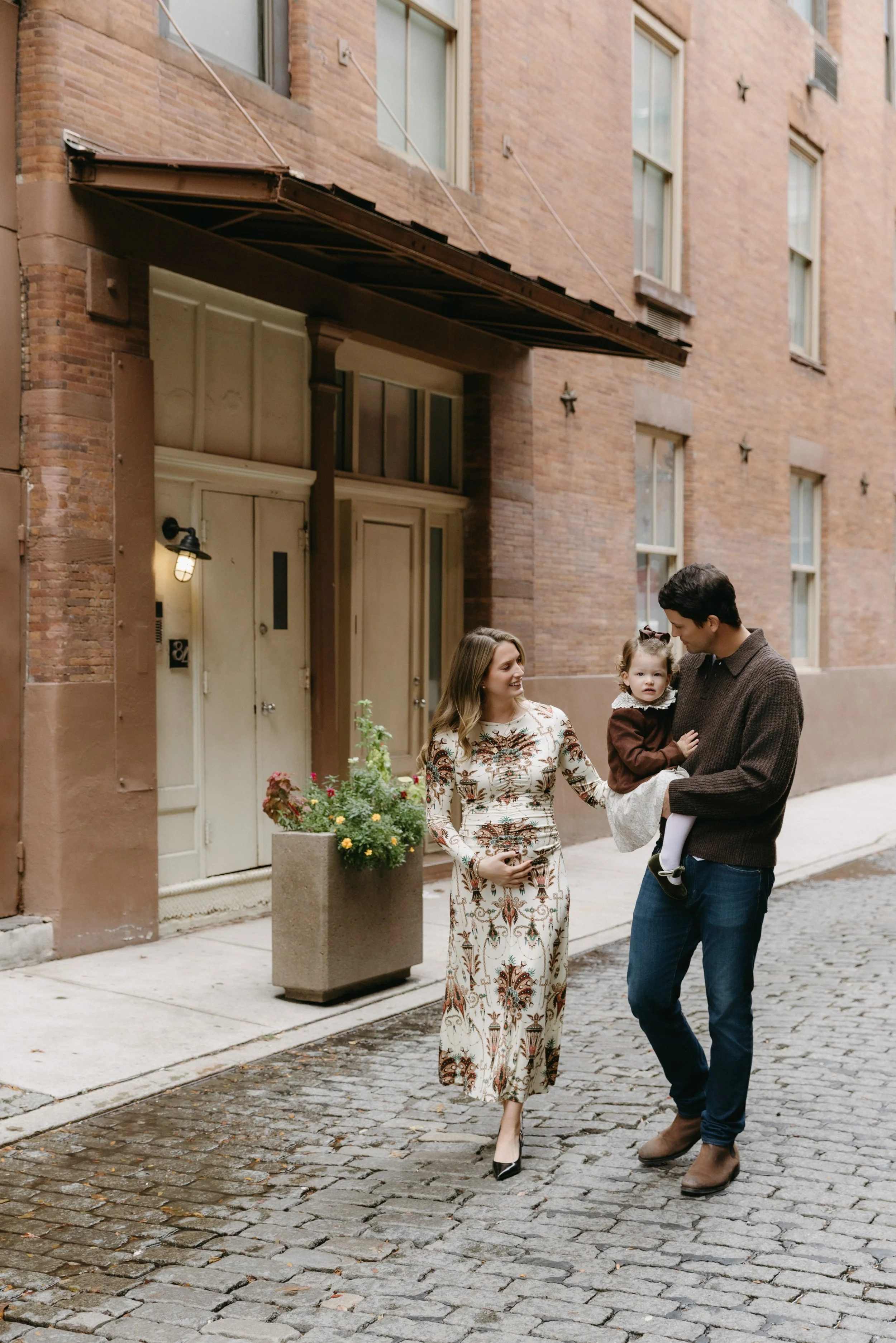 A family photograph of a family walking on a cobblestone street in TriBeCa, with a mother in a floral dress, a father in a brown sweater, and a young daughter in a brown jacket in his arms, by Maison Mancel.