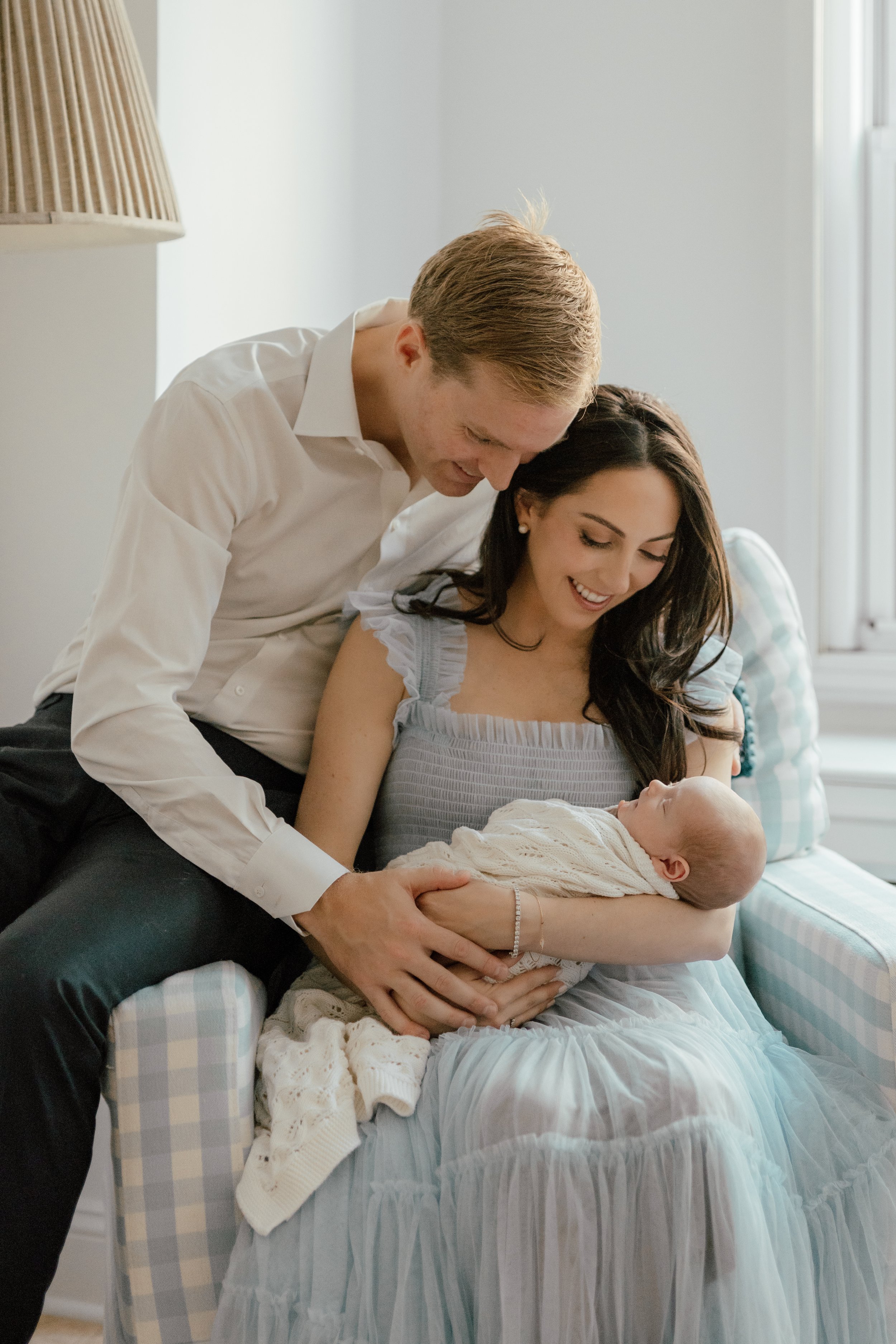 In-home newborn portrait of a mother sitting on a plaid armchair, holding a newborn baby wrapped in a cream blanket. The father is leaning over, looking at baby, in a softly lit room near a window, photographed by Maison Mancel in NYC.
