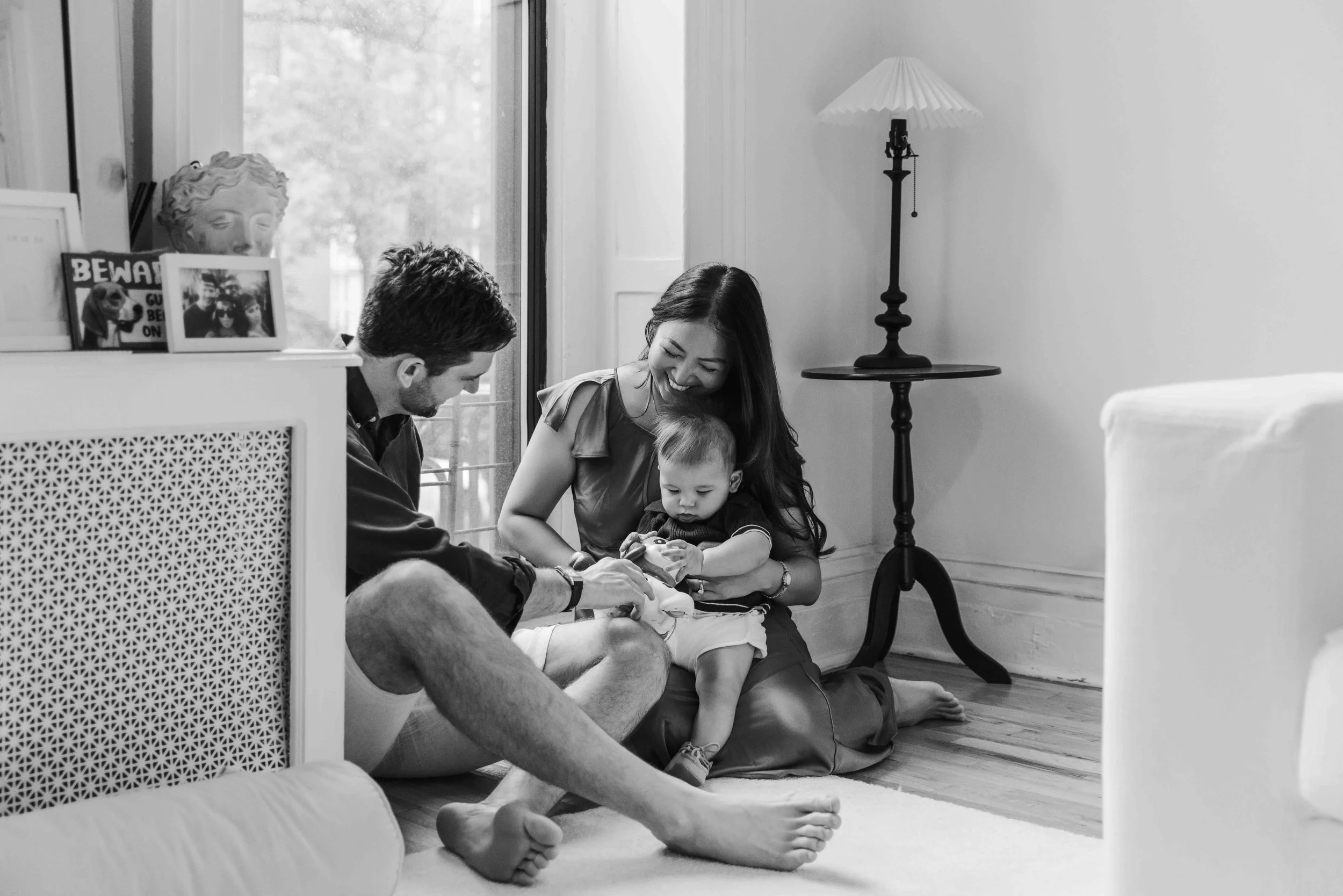 Family photograph of a family of three sitting on the floor of a living room, smiling and looking at a small object, with a window and a lamp in the background in Brooklyn - Maison Mancel.