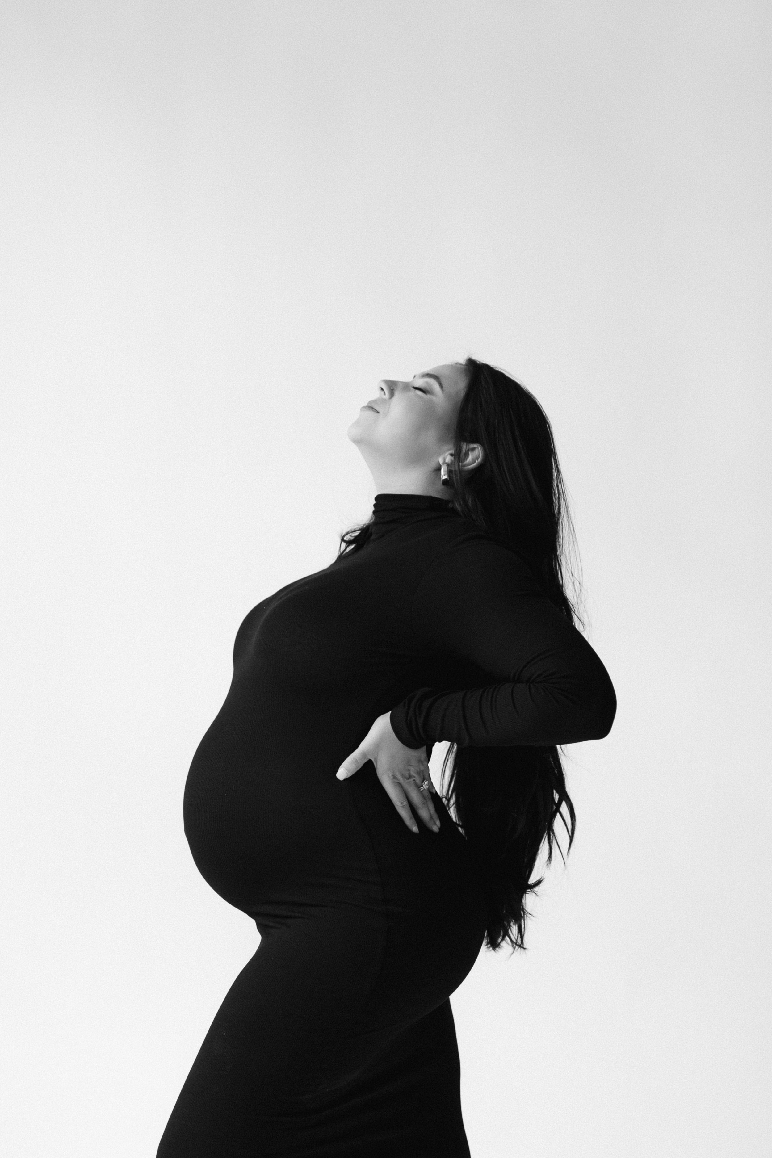 A maternity portrait of a pregnant woman with long dark hair wears a black long-sleeve dress, standing against a plain background, with her eyes closed, smiling, and one hand on her hip. Photographed by Maison Mancel in NYC.