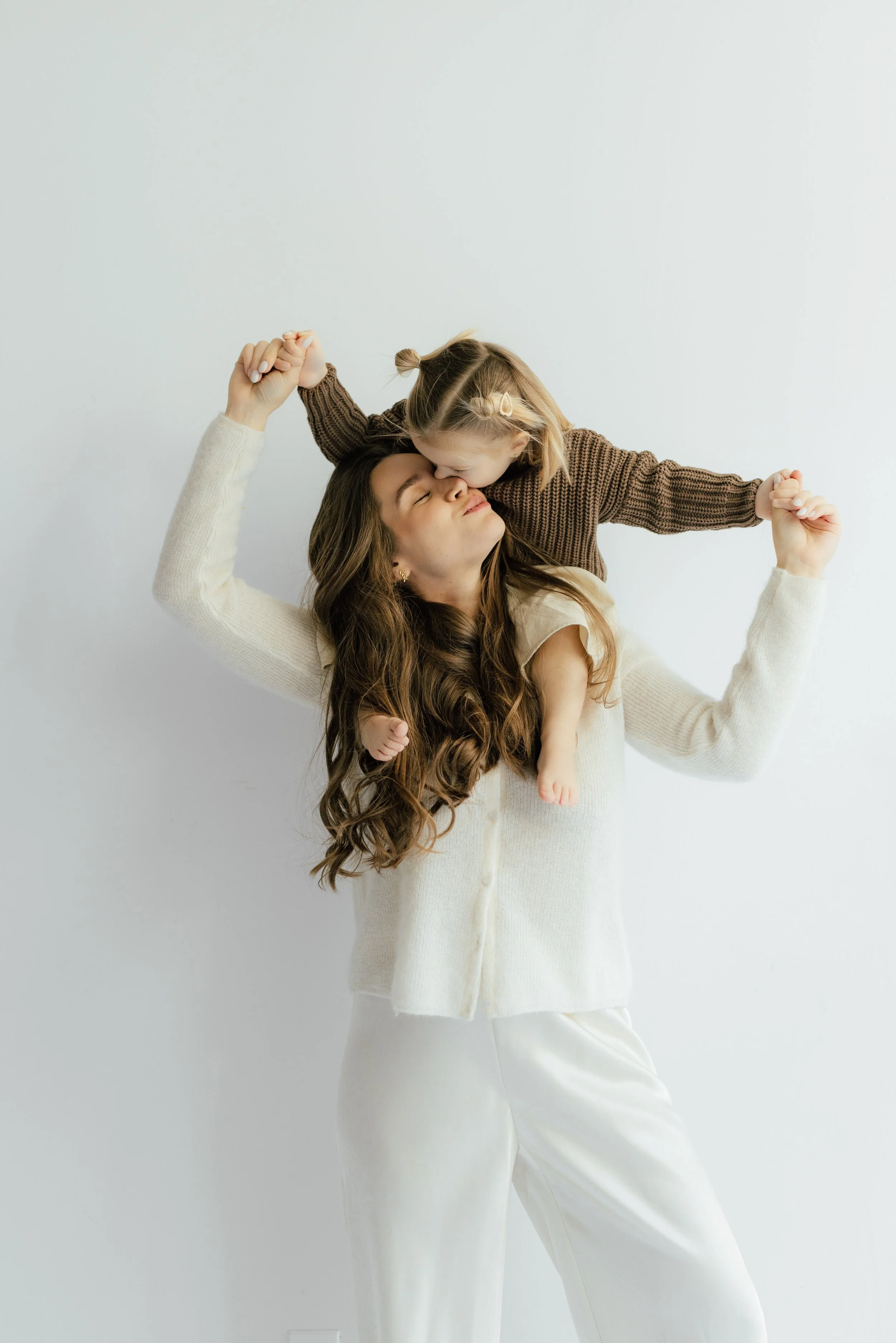 A Brooklyn studio portrait of a mother with long brown hair and a young girl with blonde hair styled in small ponytails, with the mother smiling and the girl leaning in to kiss her on the nose, both holding hands in a playful pose, by Maison Mancel.