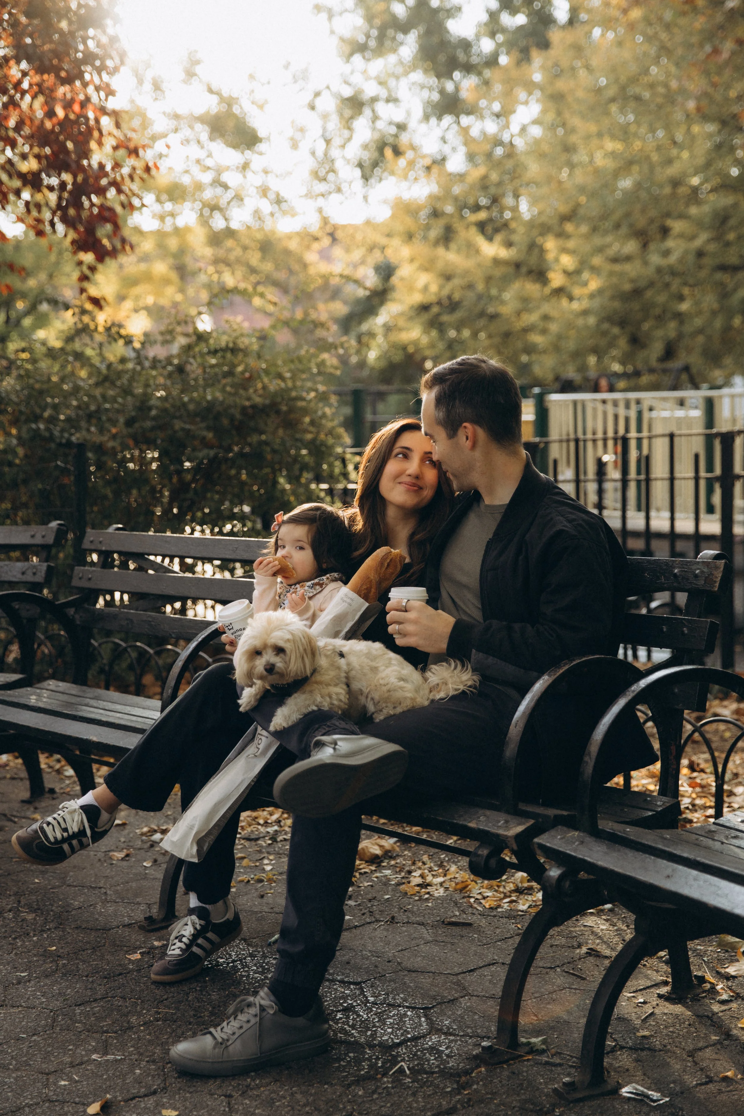 A portrait of a family of three with a small dog sitting on a Brooklyn park bench, sharing coffee and snacks during autumn, with trees and fallen leaves in the background, photographed by Maison Mancel.