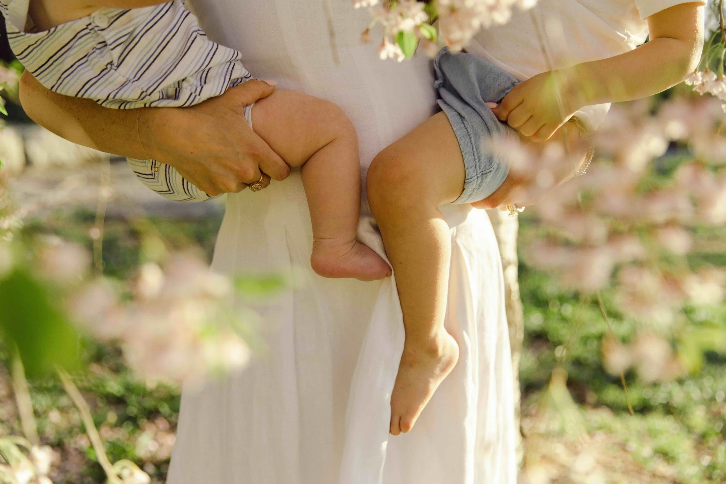 A family portrait of a mother holding her children outdoors near pink flowering trees in Brooklyn Heights, by Maison Mancel.