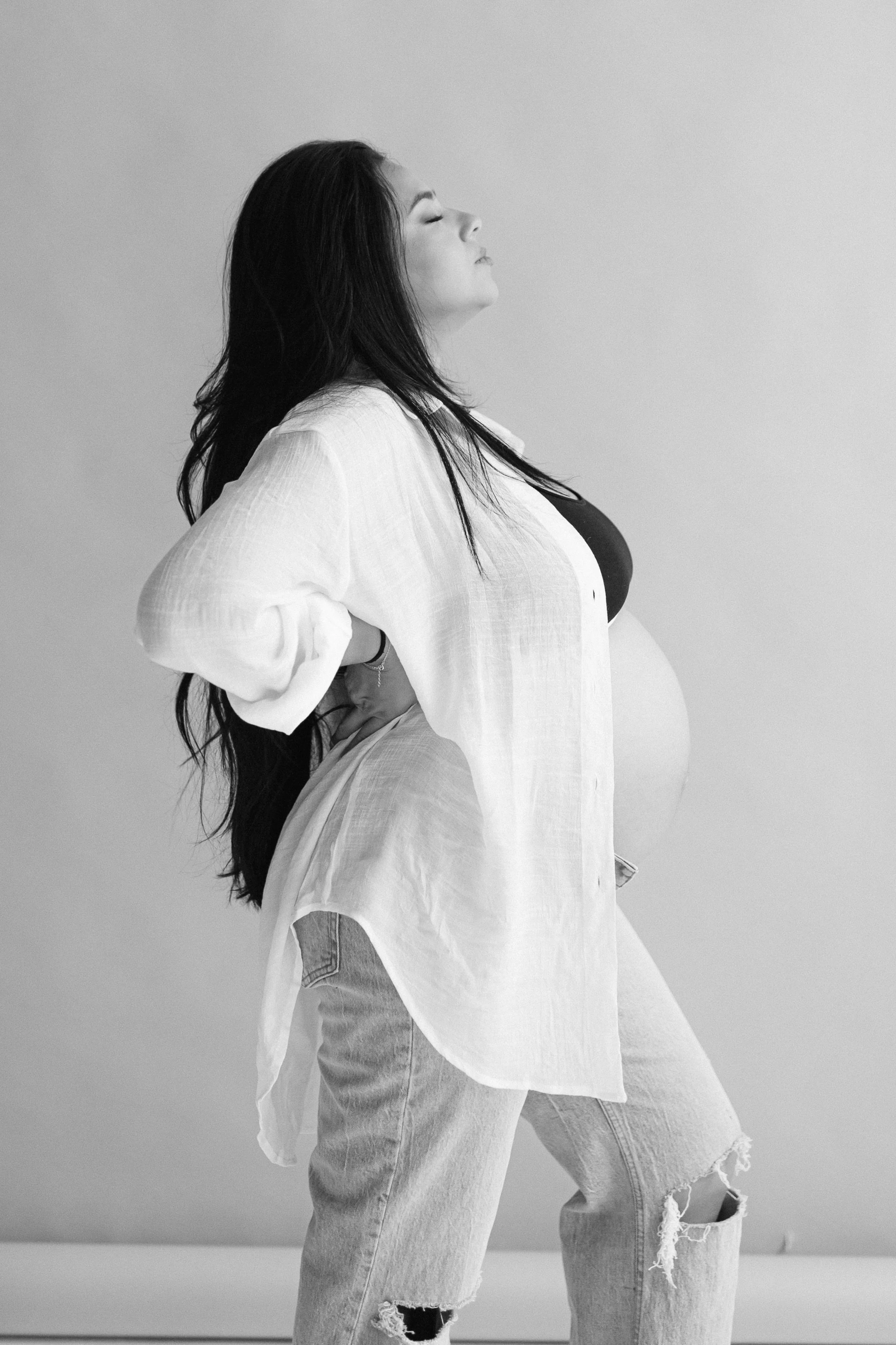 Black and white maternity photo of a pregnant woman standing sideways with hands on her hips, wearing a black top, white button-up shirt, and ripped jeans, with her eyes closed and her head tilted slightly back. Photographed by Maison Mancel in NYC.