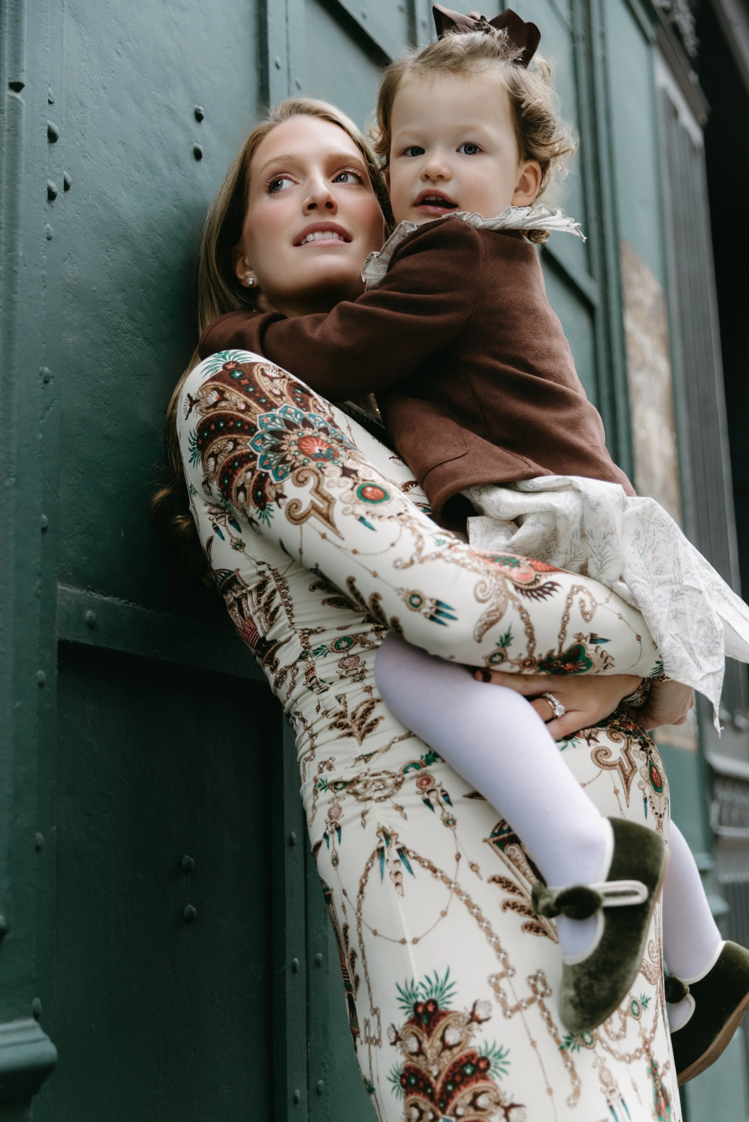 A pregnant mother with a patterned dress holds a young girl with a brown jacket and white tights, against a green wall in TriBeCa, captured by Maison Mancel.