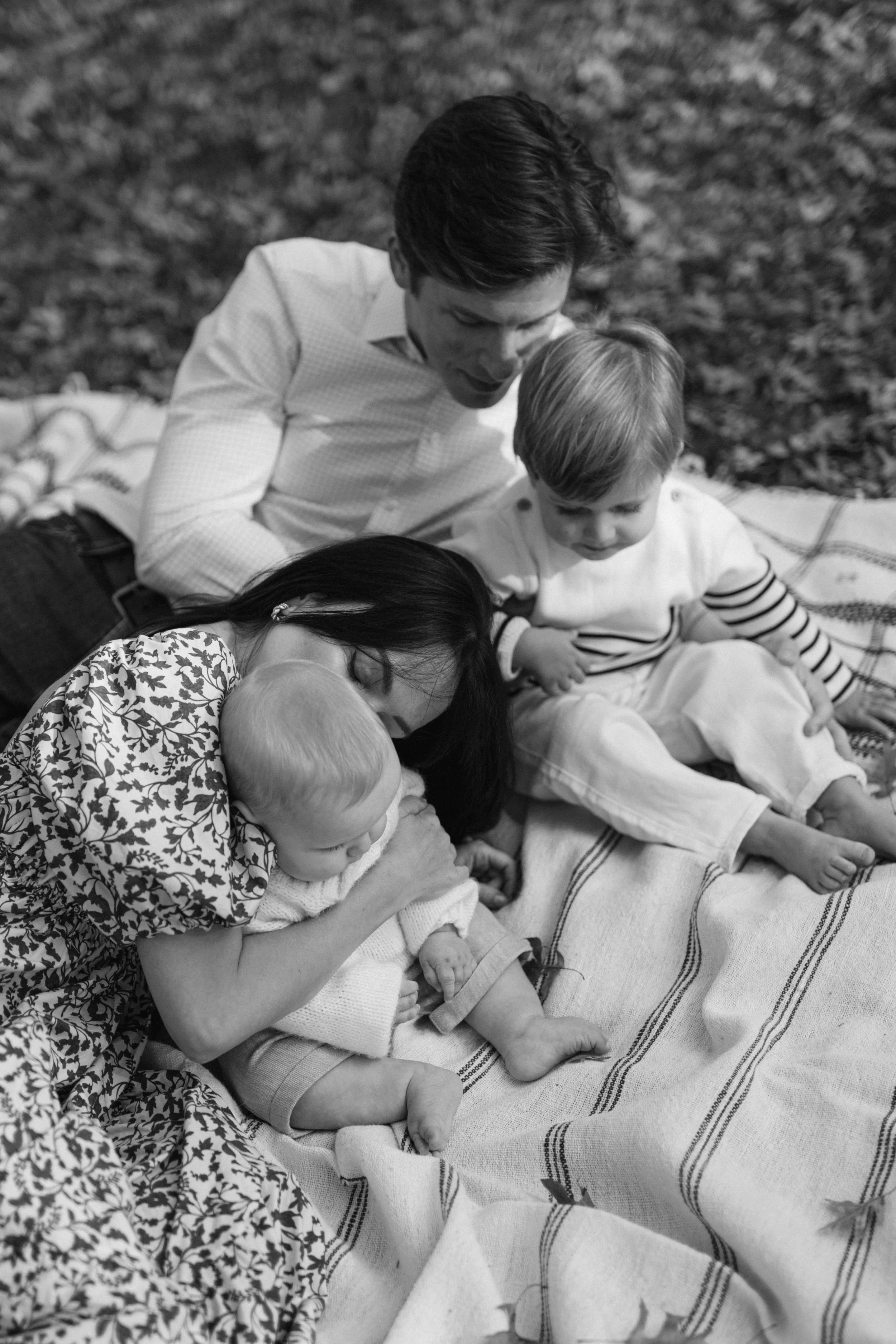 A lifestyle portrait of a family of four lying together on a blanket outdoors, including two adults and two children, with the youngest child being held by one of the adults, in Central Park, photographed by Maison Mancel.
