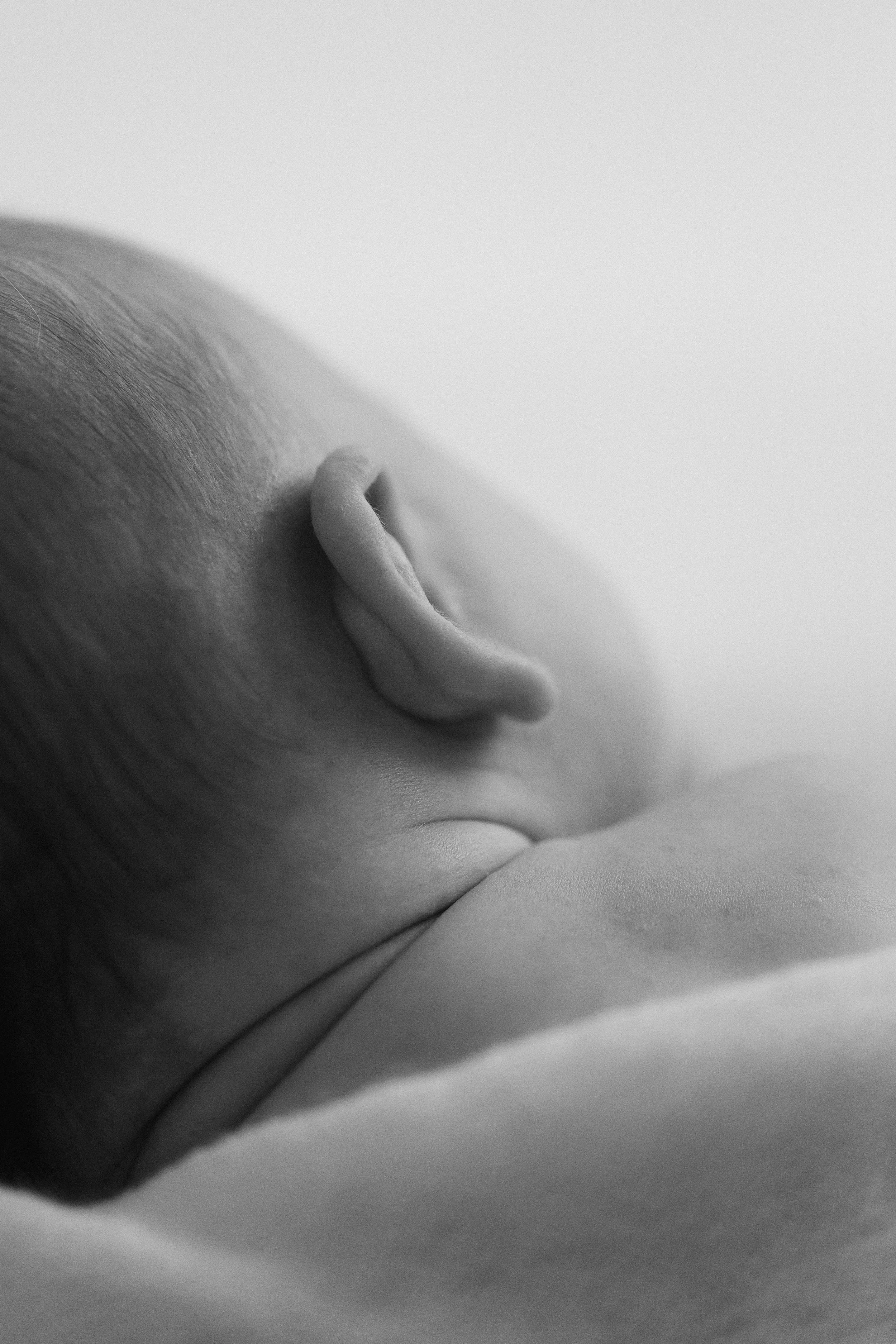 Black and white close-up of a newborn's head, focusing on the ear and part of the face, resting on a soft surface. Photographed by Maison Mancel in NYC.