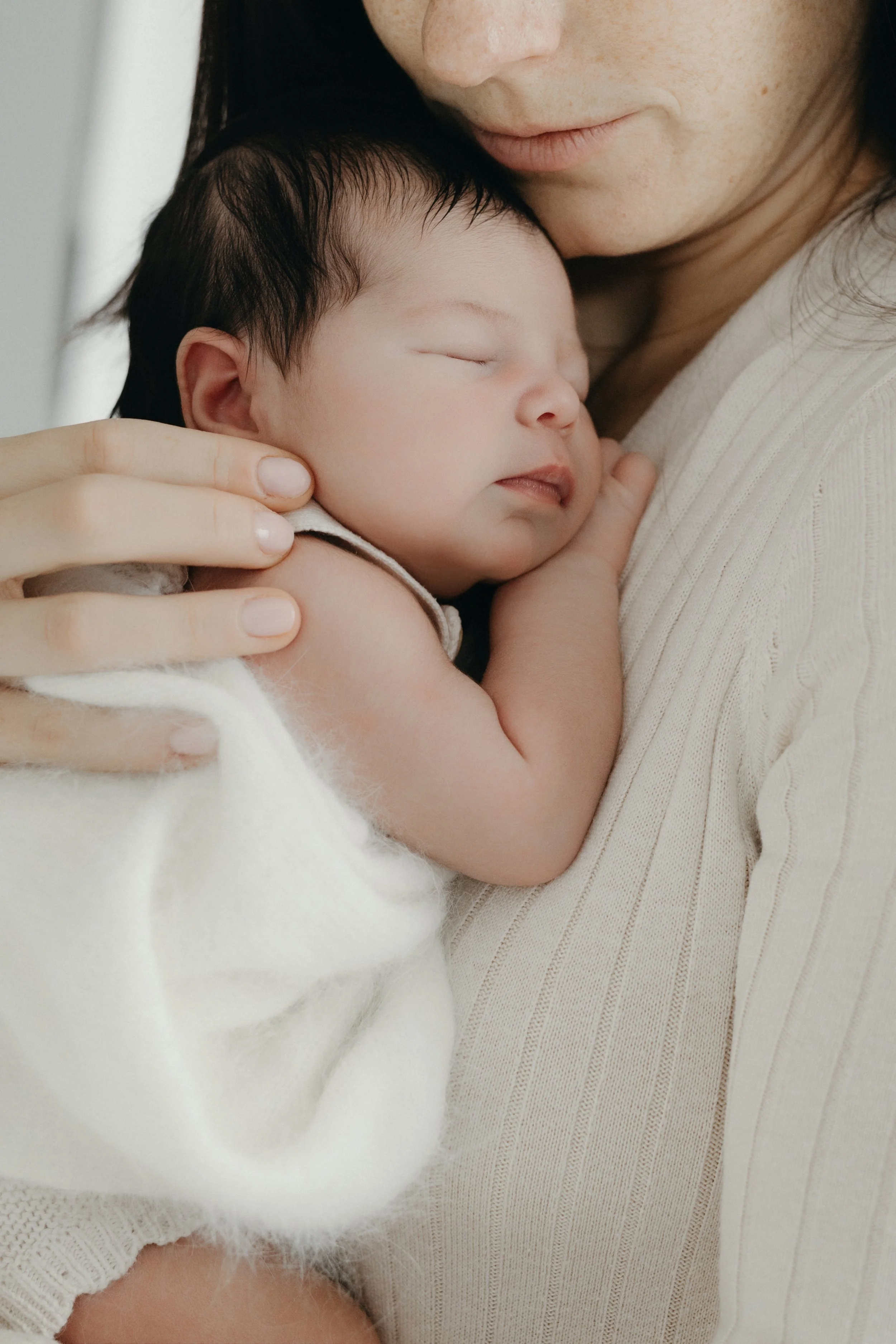 Portrait close-up of a mother holding a sleeping baby with dark hair, gently cradling the baby's head with one hand, both wearing light-colored clothing, photographed by Maison Mancel in-studio in NYC.