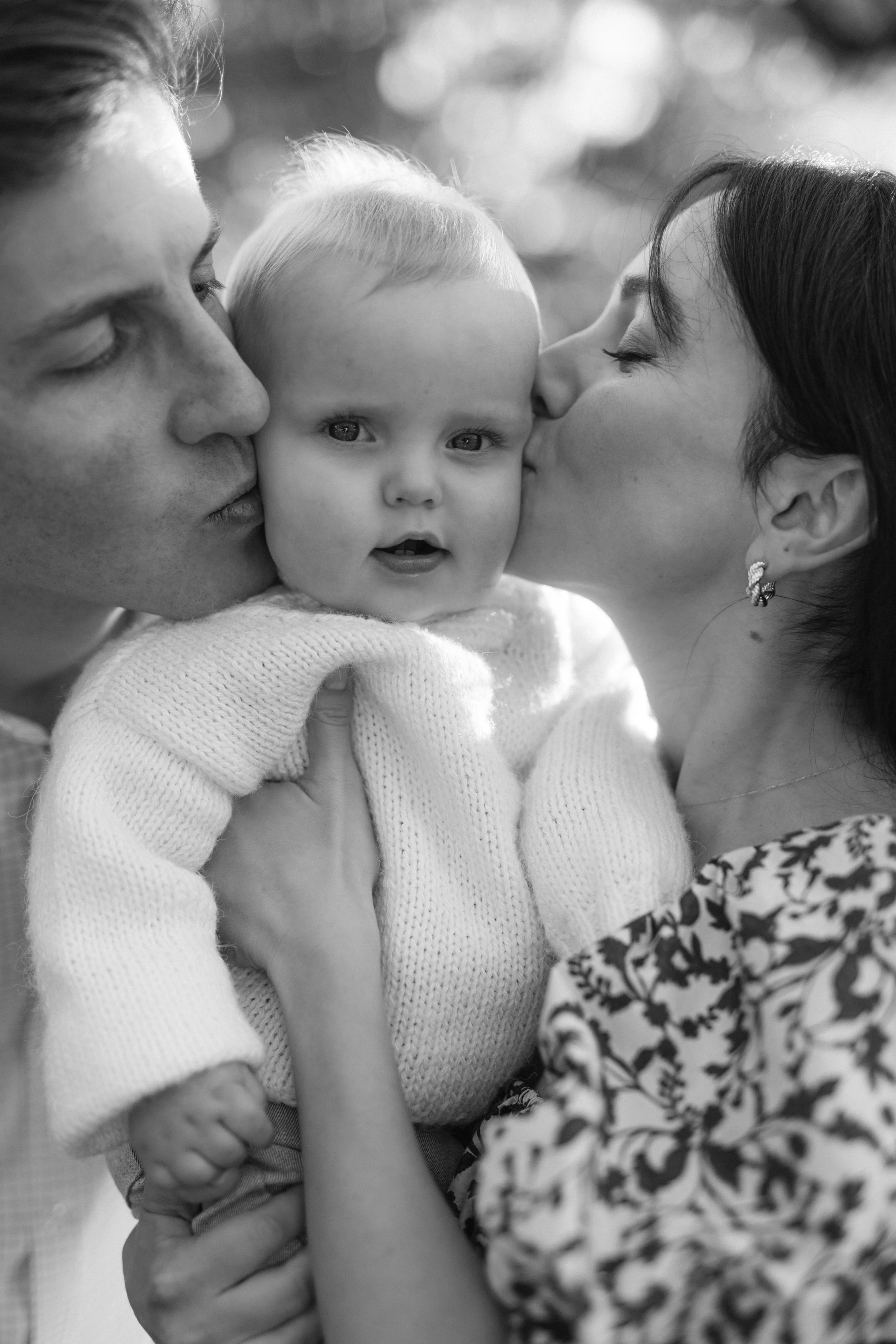 Portrait of a child being kissed on the cheeks by  parents, in Central Park. The mother is wearing earrings and a patterned top, and the man is in a collared shirt. The child is looking at the camera with a neutral expression. By Maison Mancel.