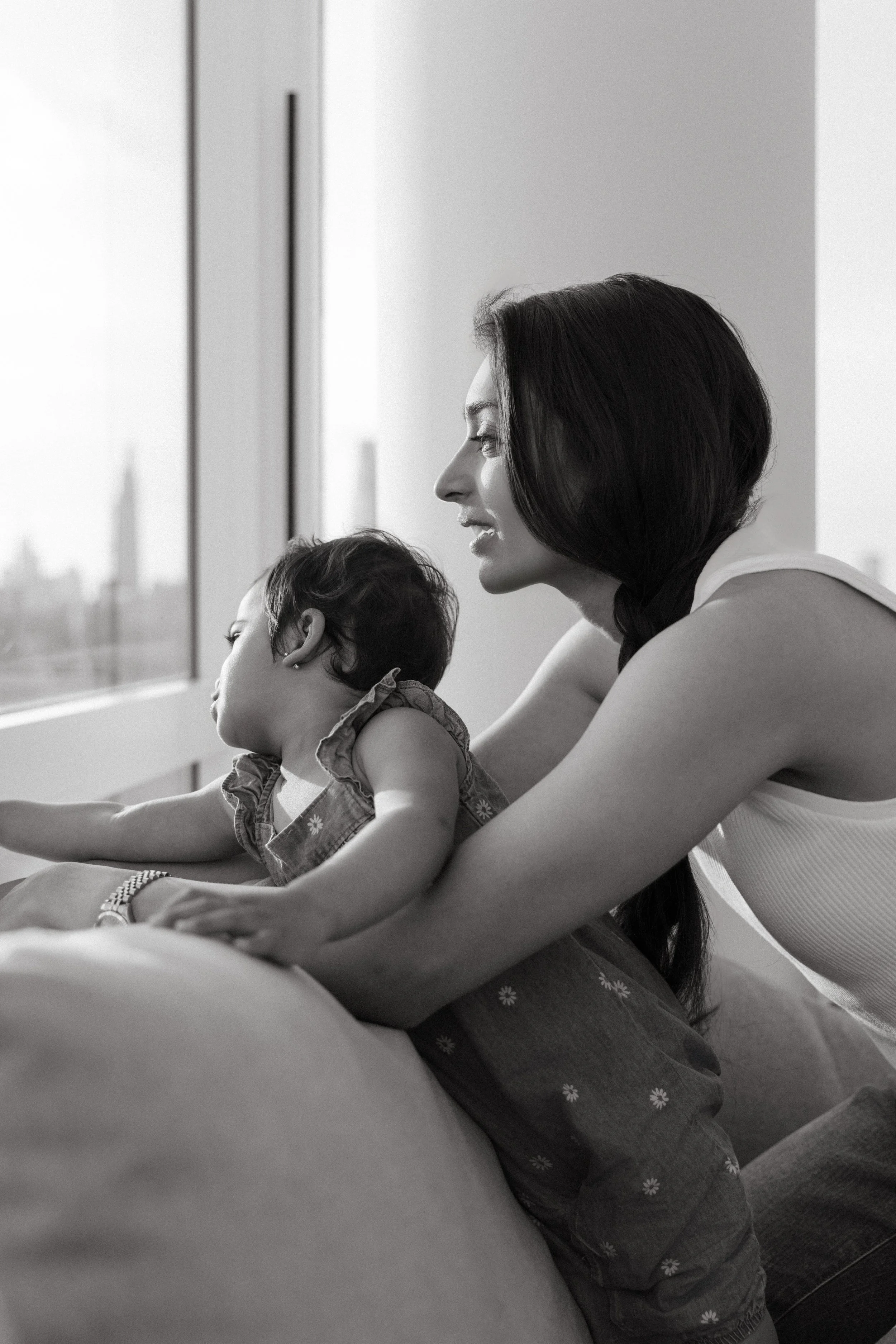 A family portrait showing a mother with dark hair and a sleeveless top leaning on a couch, holding a young child with curly hair and patterned clothing, looking out a large window during daytime, in-home in Queens by Maison Mancel.