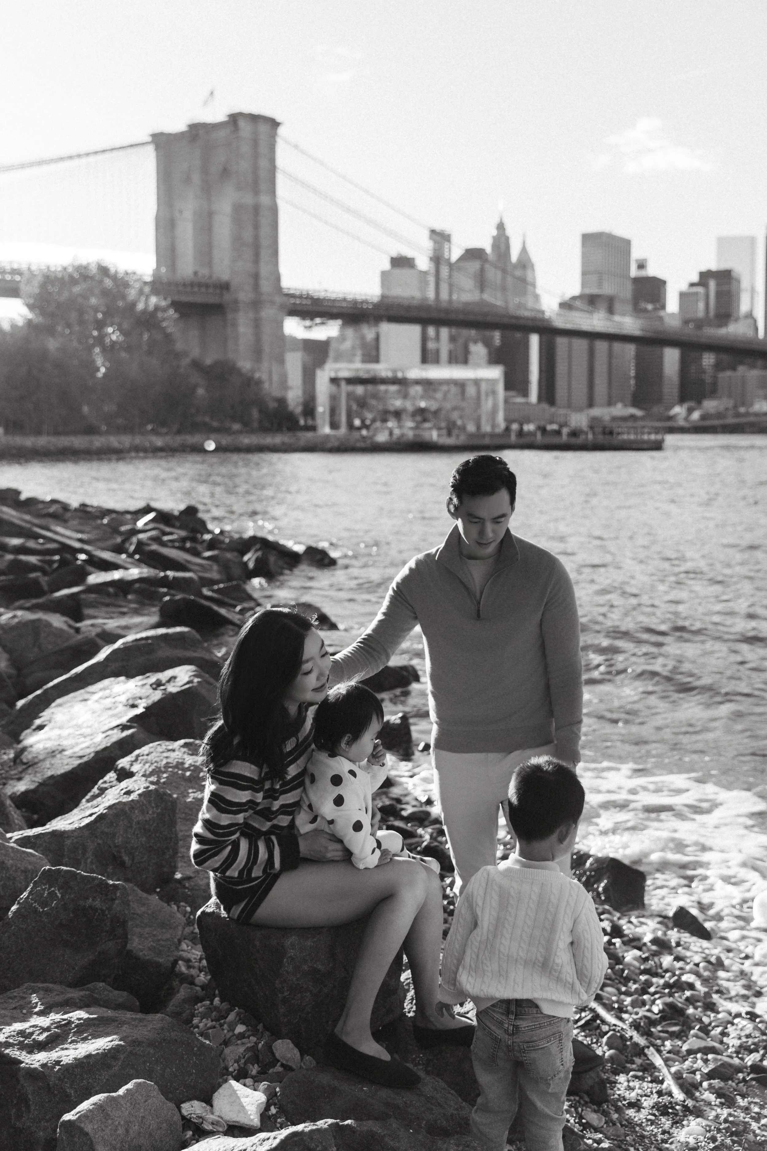 A family of four sitting and standing on rocks by the water in New York City with Brooklyn Bridge and Manhattan skyline in the background, black and white photo, captured by Maison Mancel.