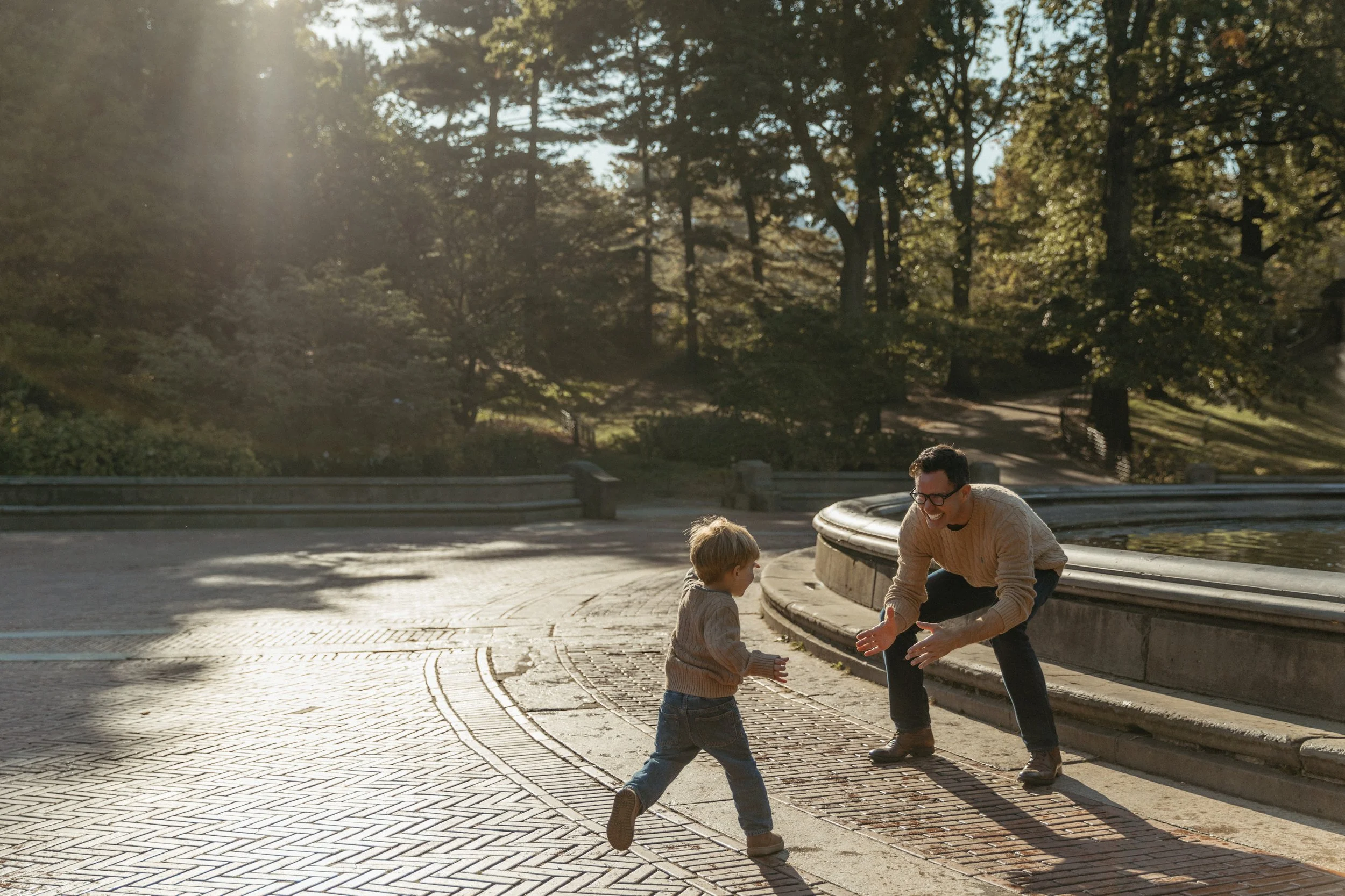 A portrait of a father and a young boy playing and laughing together by a fountain in a park, surrounded by trees and sunlight filtering through the leaves in Central Park, photographed by Maison Mancel.
