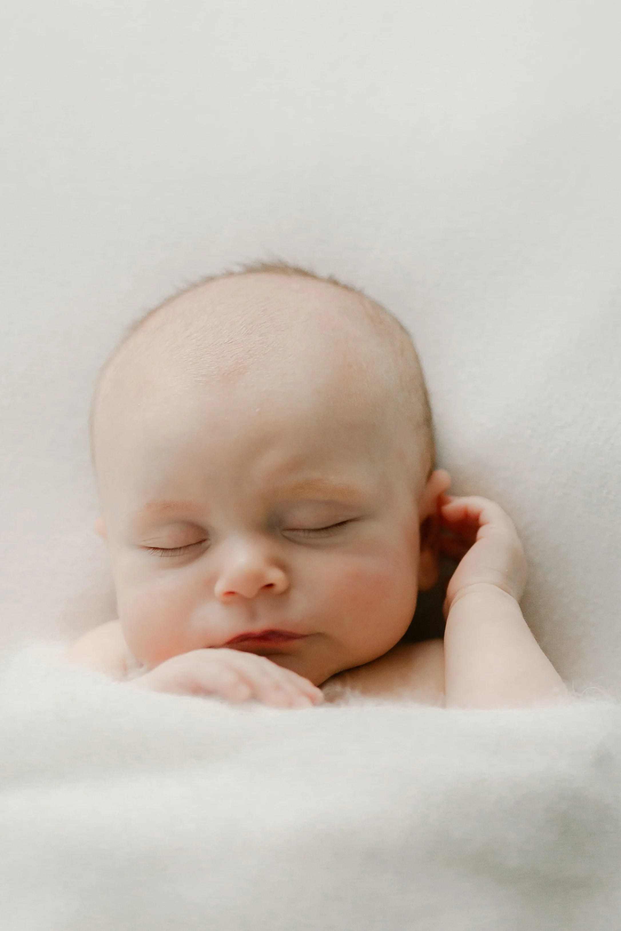 Close-up of a sleeping newborn baby with light skin, soft facial features, and a bald head, resting on a white surface with one hand near the ear. Photographed by Maison Mancel in NYC studio.