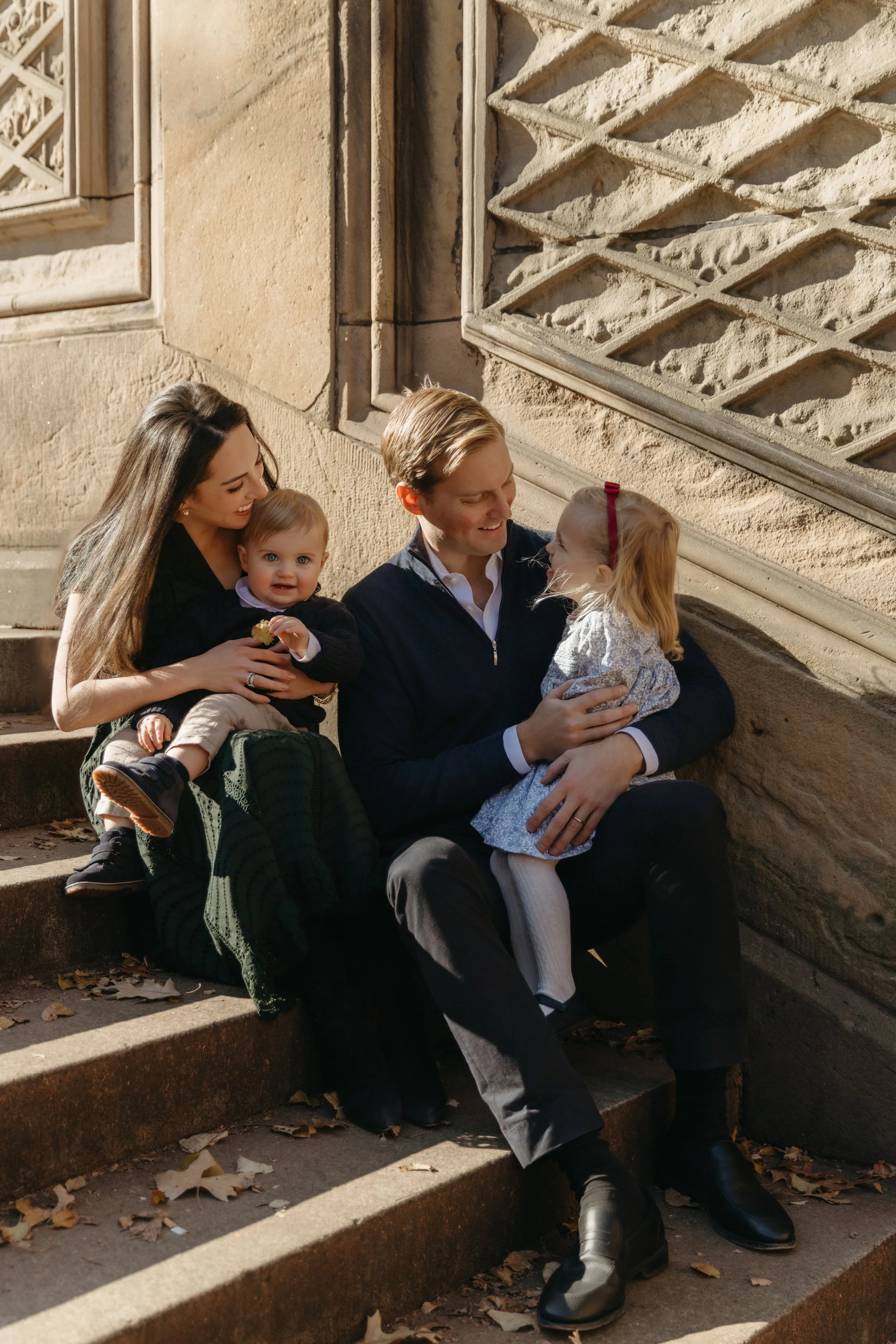 A lifestyle family portrait featuring a family of four sitting on outdoor steps in Central Park, a mother, father, and two young daughters, smiling and enjoying each other's company during fall, with leaves scattered on the steps, by Maison Mancel.