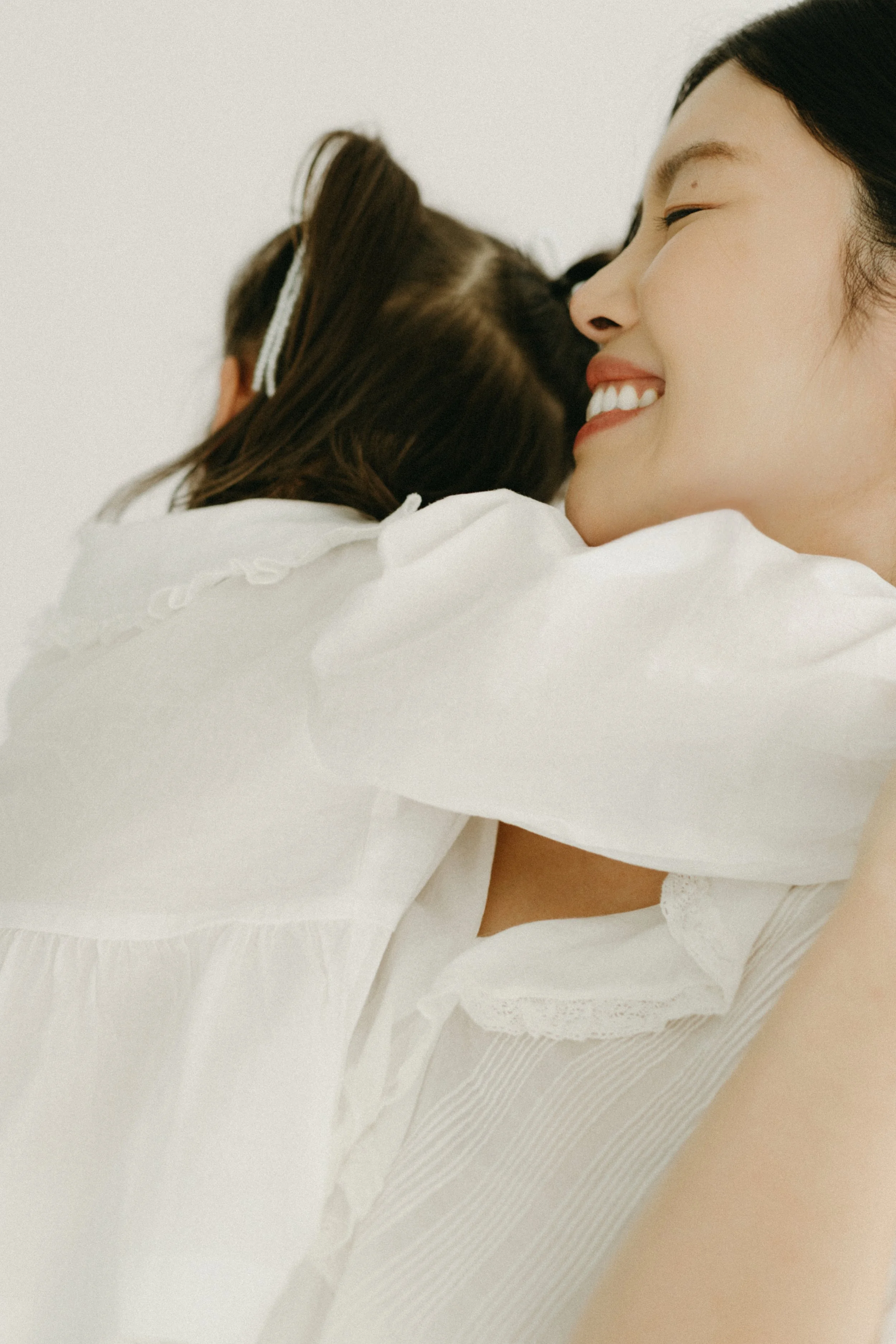A  family portrait of a mother with dark hair smiling and hugging a young girl with brown hair and a hair clip, in-studio, photographed by Maison Mancel.