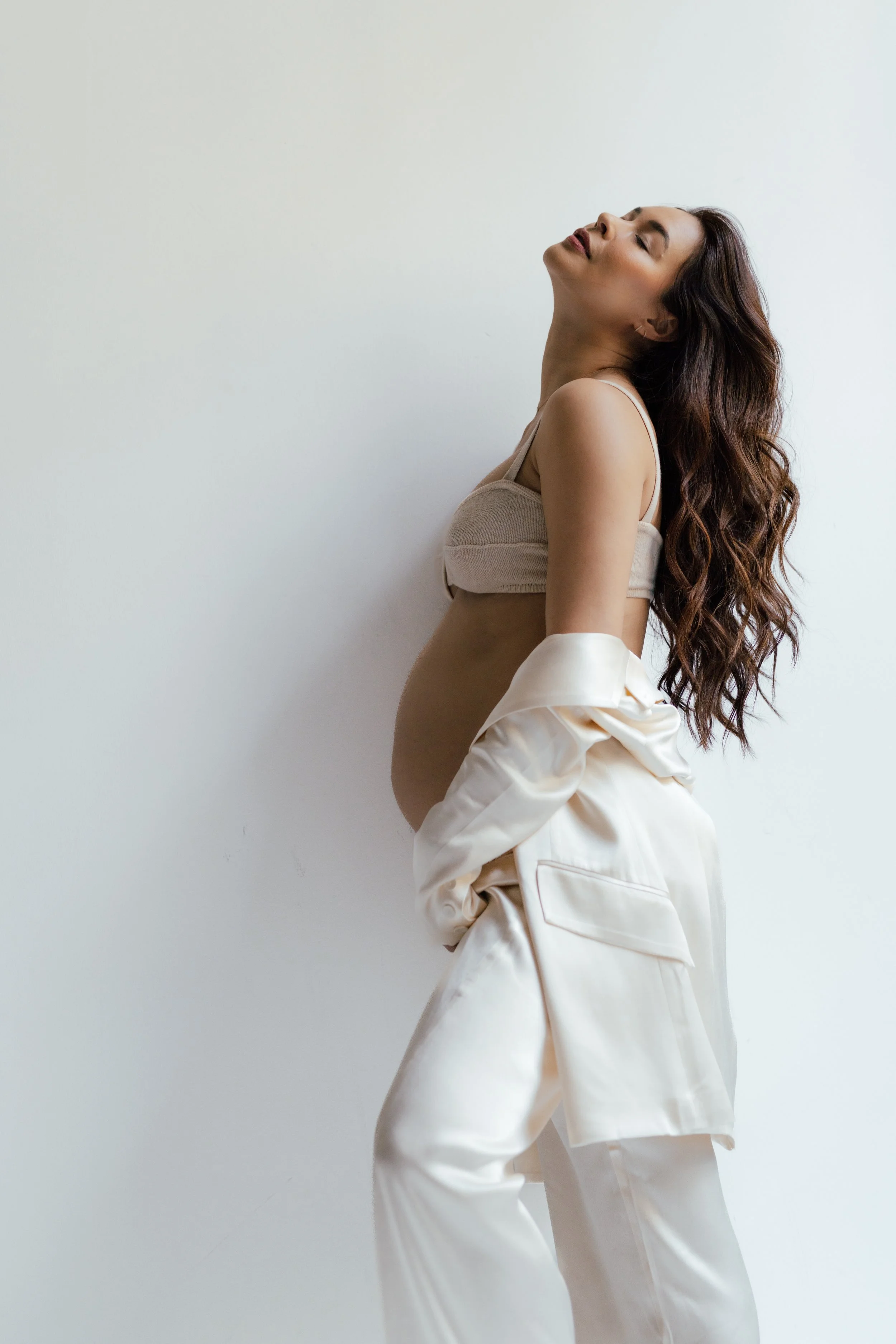 A pregnant woman with long, wavy brown hair, wearing a beige crop top and cream-colored satin pajamas, standing against a plain white wall with her eyes closed and head tilted back. Photographed by Maison Mancel in Industry City, Brooklyn, NYC.