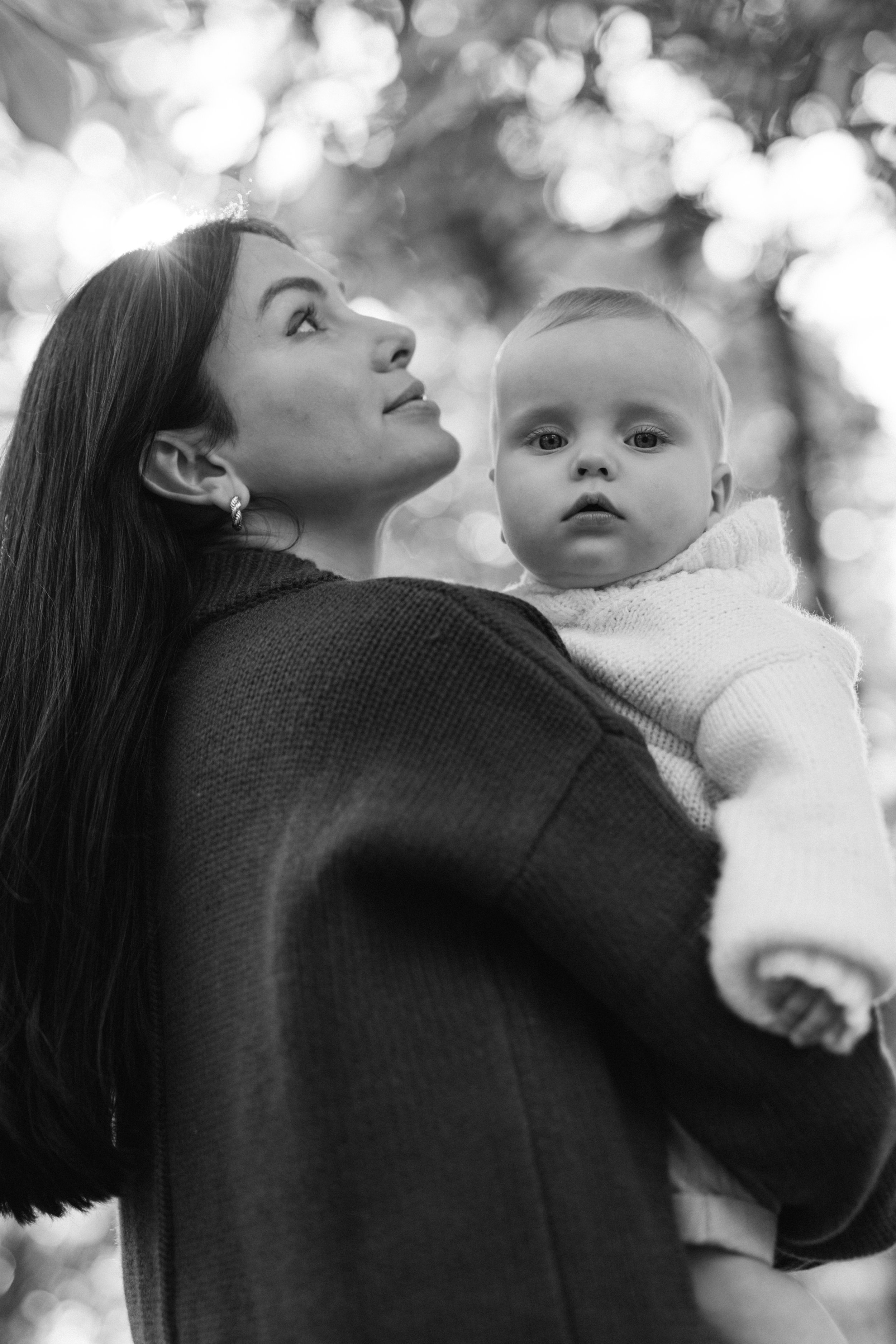 A motherhood portrait of a mother holding a young child outdoors with blurred trees and sunlight in the background in Central Park, photographed by Maison Mancel.