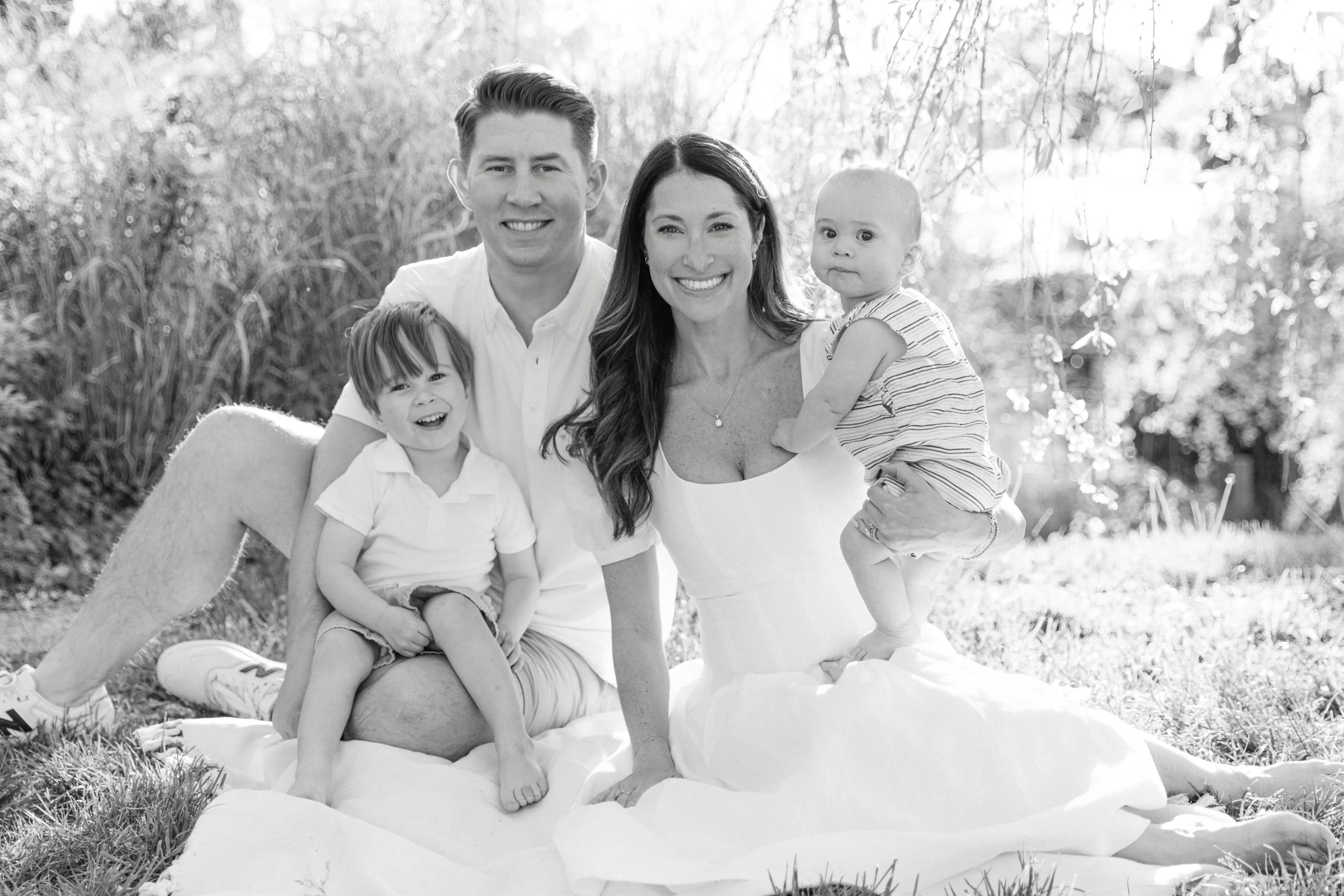 A family portrait showing a family of four sitting on a blanket outdoors in Brooklyn Heights, smiling at the camera, with trees in the background, by Maison Mancel.