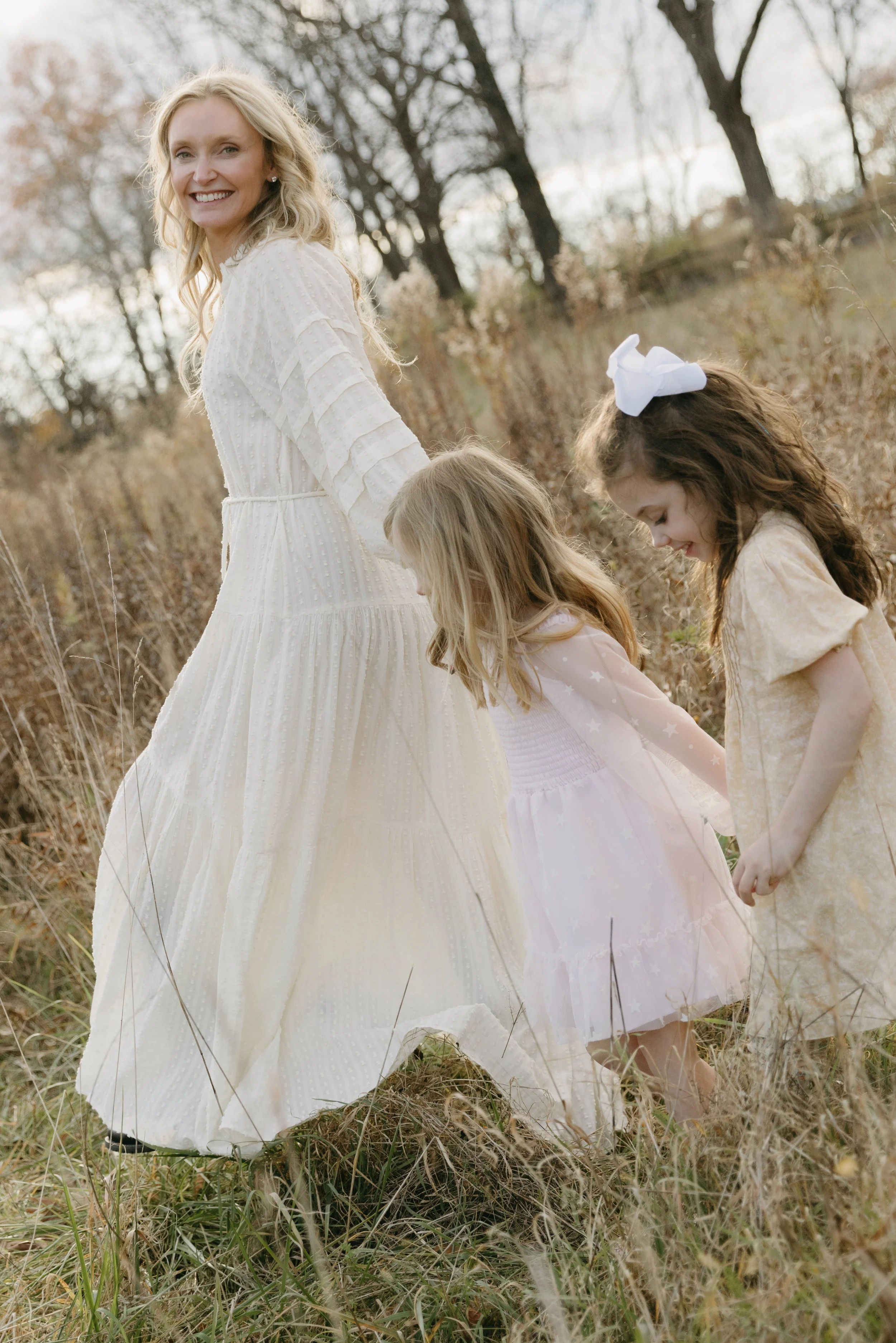 A family portrait of a smiling mother in a white dress walking outdoors with two young girls in pastel dresses, holding hands in a grassy field with trees in the background during fall, photographed by Maison Mancel in New Jersey.
