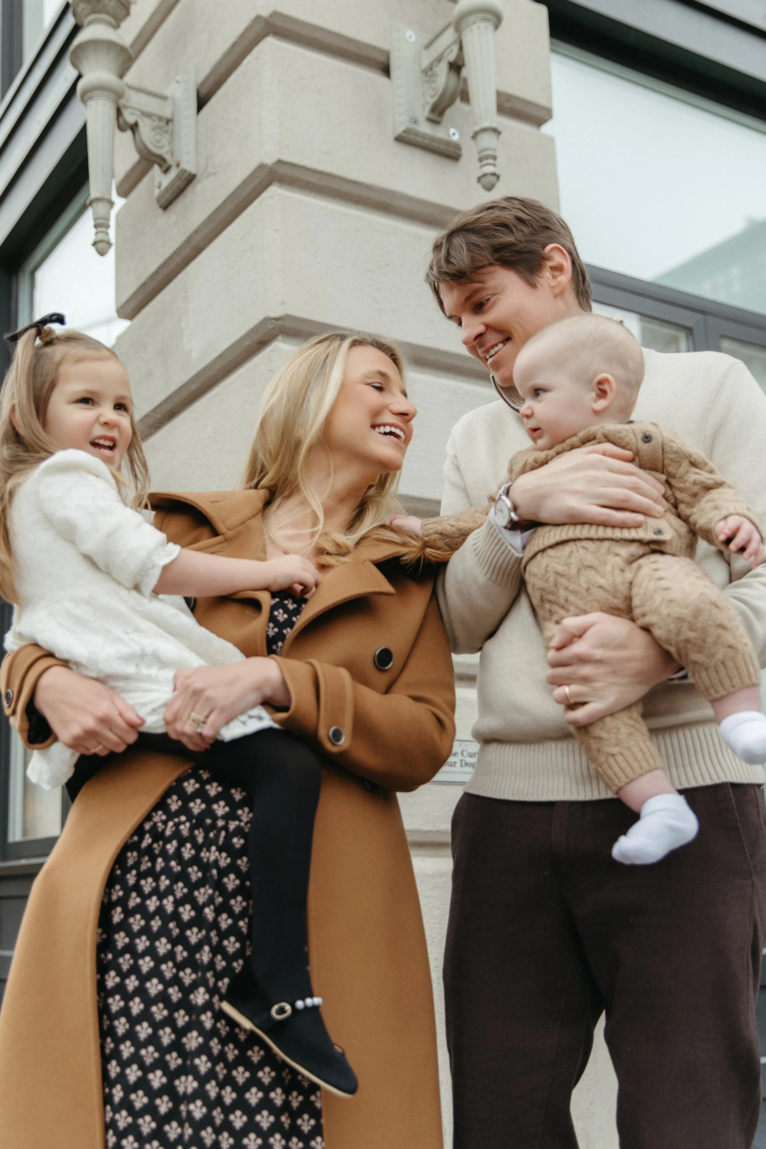An outdoor lifestyle portrait of a family of four standing outside a building, smiling and interacting. The mother is holding a young girl, and the father is holding a baby. All are dressed in warm clothing. Taken in Dumbo by Maison Mancel.