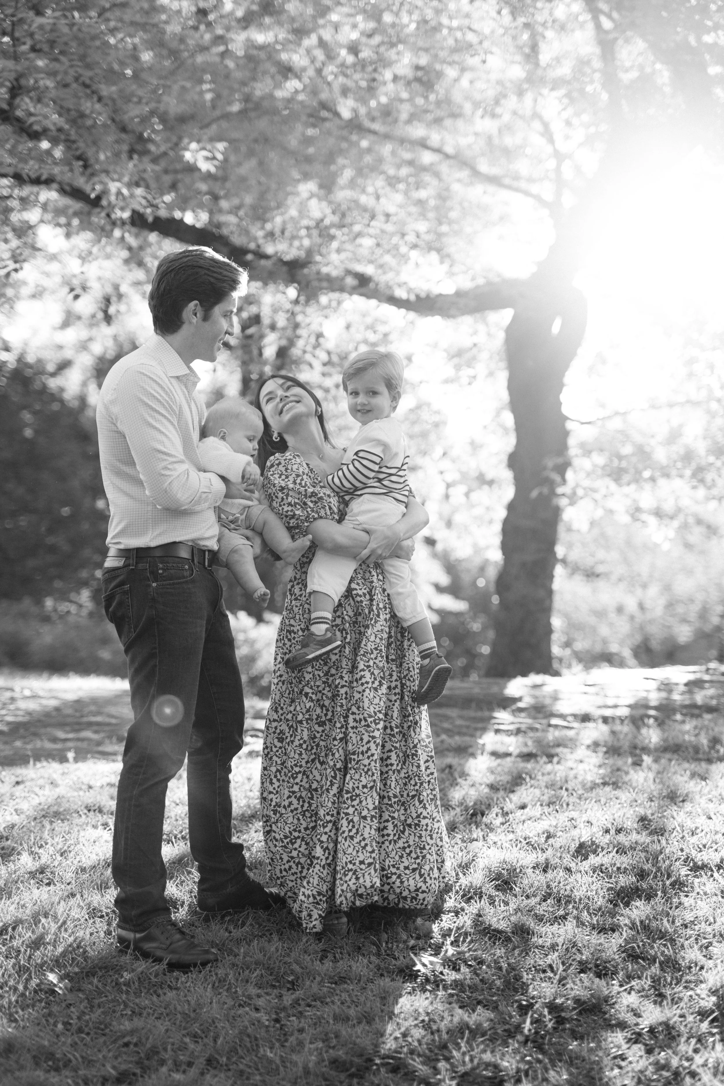A portrait of a family of four enjoying time outdoors, with the parents holding two young children, and the sun shining through trees in the background in Central Park, by Maison Mancel.