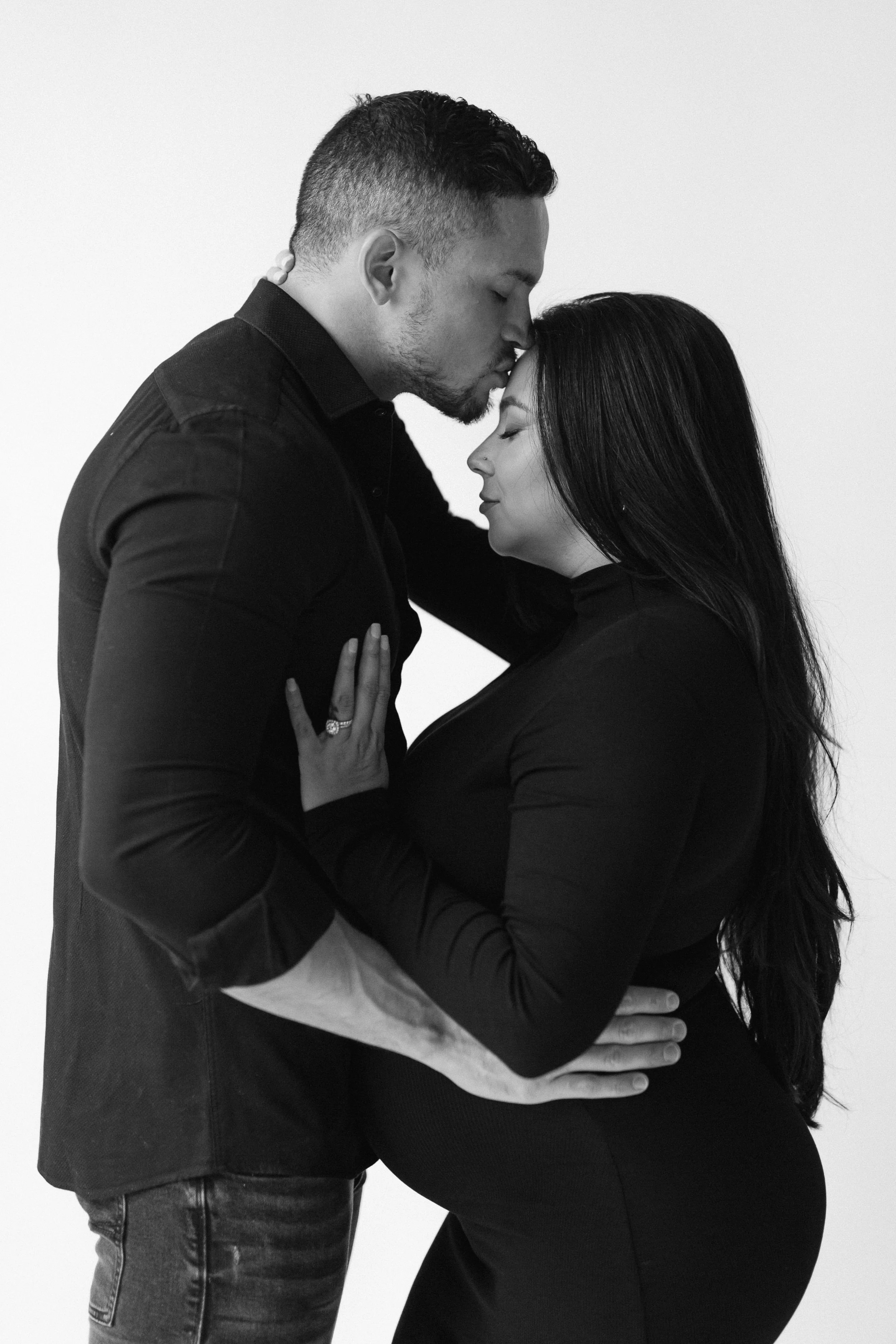A black and white maternity portrait of a man and pregnant woman in an intimate embrace, with the man gently kissing the woman's forehead and both with eyes closed. Photographed by Maison Mancel in-studio in Brooklyn.