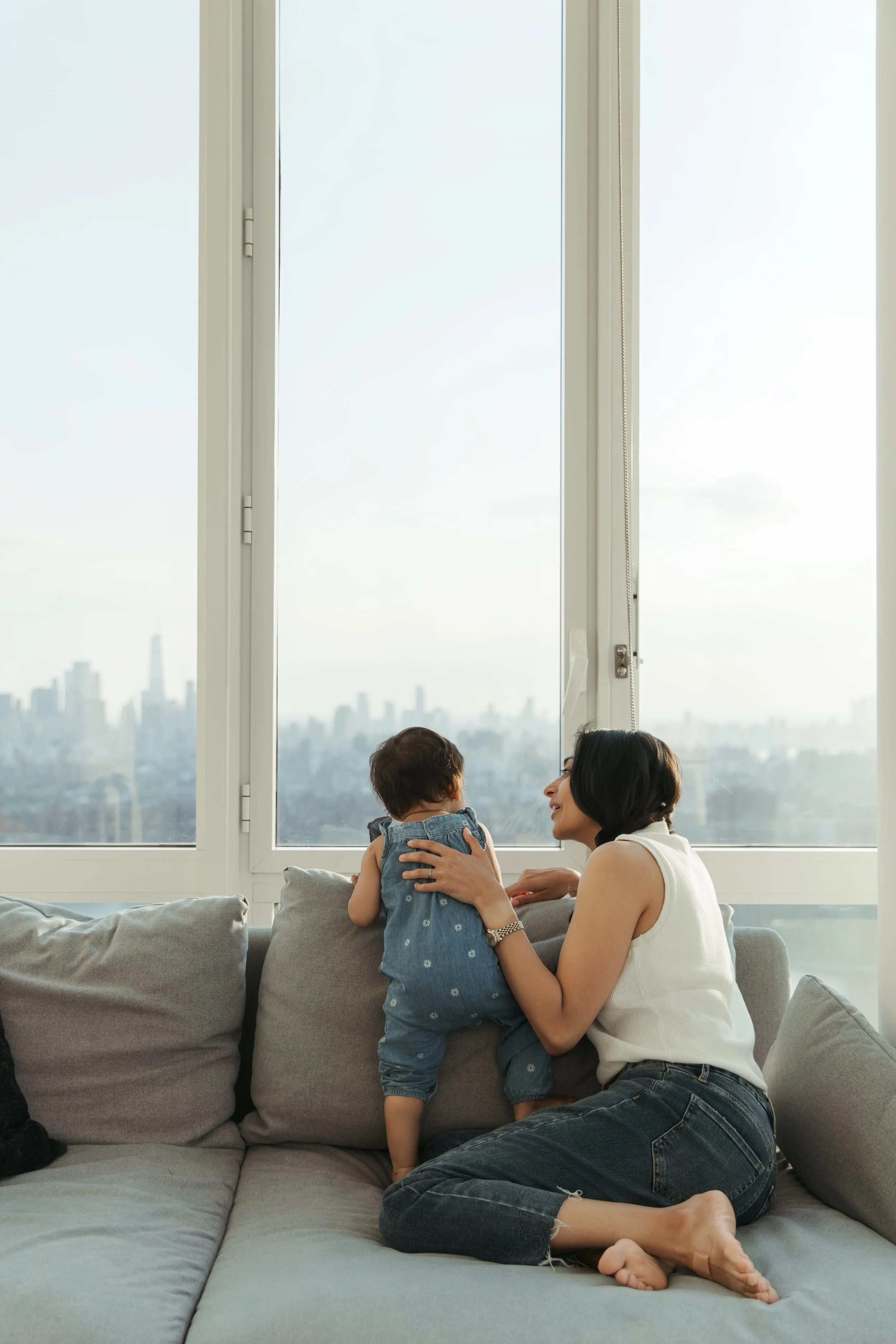 A family portrait of a mother  and a young child sit on a gray couch near a large window with a city skyline view, engaging in a playful and affectionate moment, photographed by Maison Mancel in NYC.