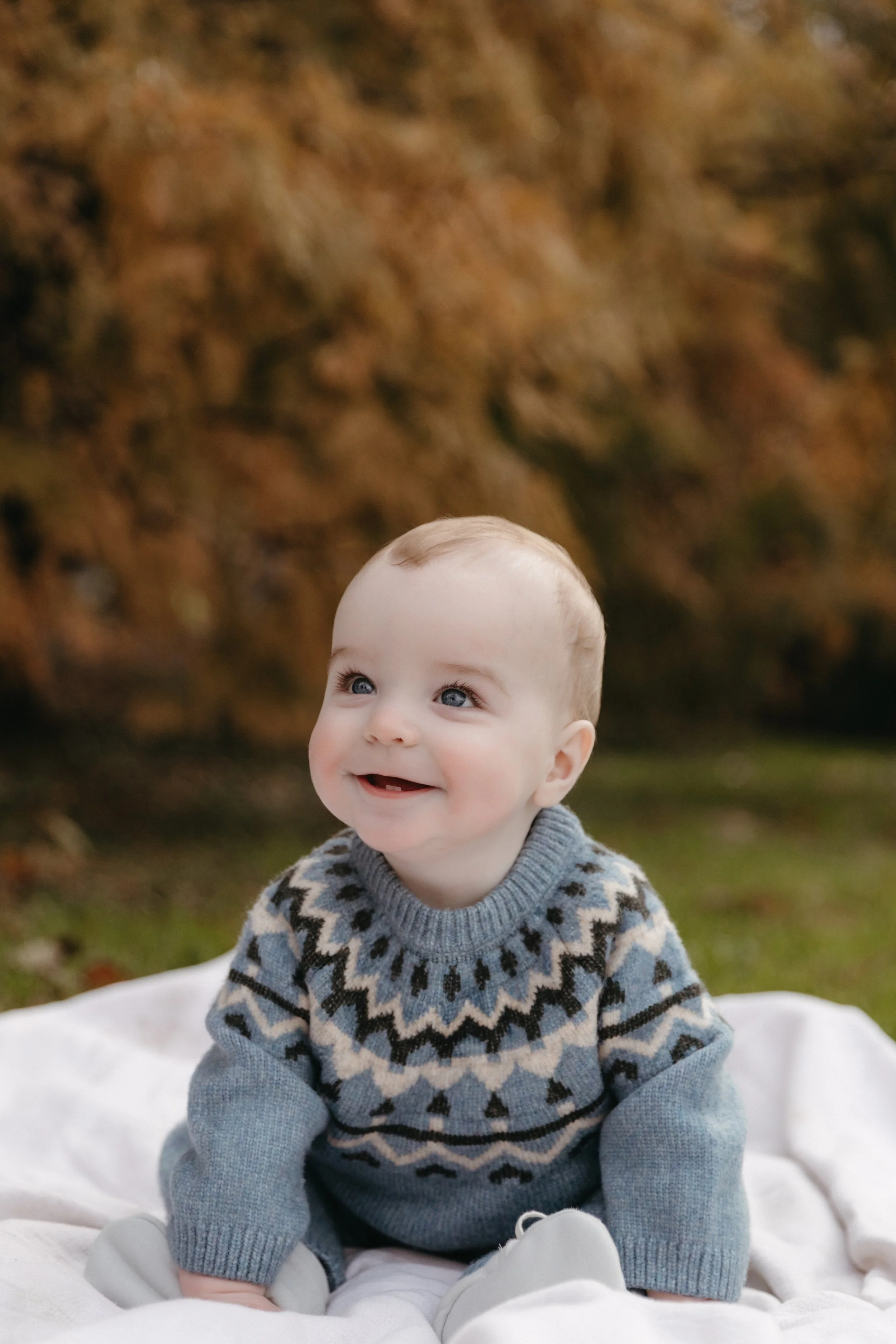 A family portrait featuring a smiling baby with blue eyes and a few teeth, wearing a patterned gray and black sweater, sitting outdoors in NYC on a white blanket with autumn trees in the background - Maison Mancel.