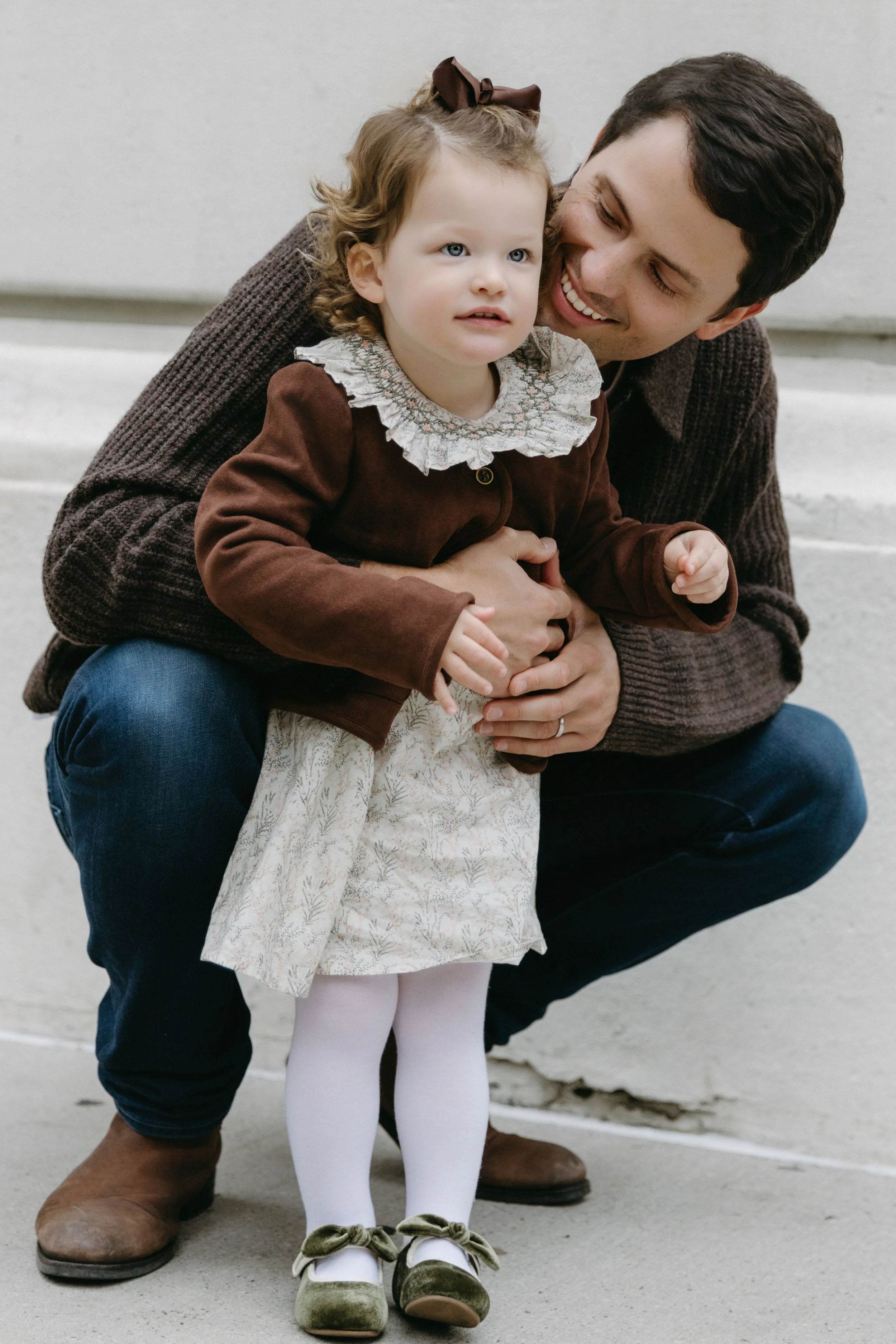 A photograph of a young girl with curly hair, dressed in vintage-style clothing, stands in front of a smiling father squatting outdoors on a sidewalk in TriBeCa, holding hands with the father, by Maison Mancel.