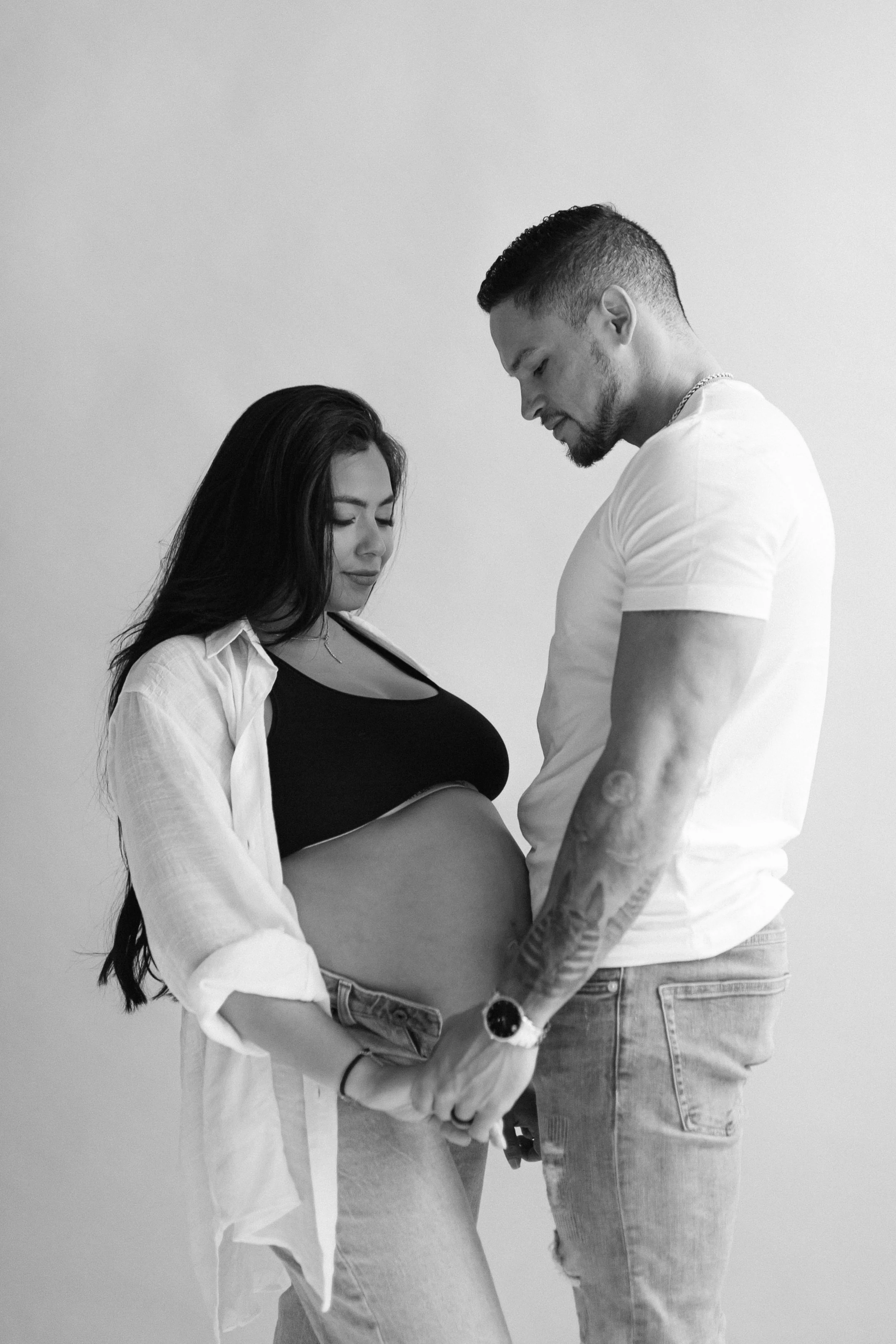 A pregnant woman and her partner holding hands, standing close with their foreheads touching, in a black and white photo.
