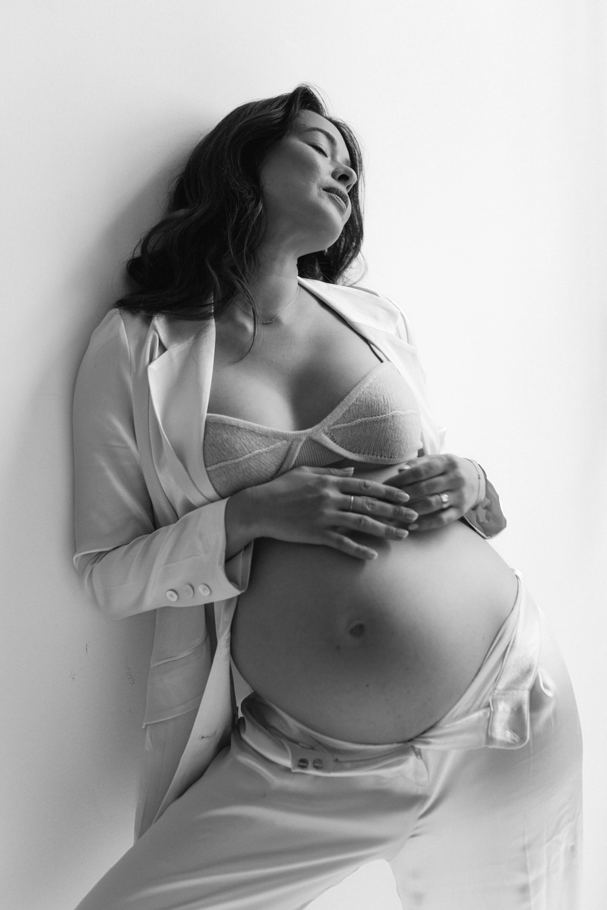 Black and white photograph of a pregnant woman with dark, wavy hair, leaning against a plain wall, wearing a bra, unbuttoned shirt, and pants, with her eyes closed and hands resting on her belly.