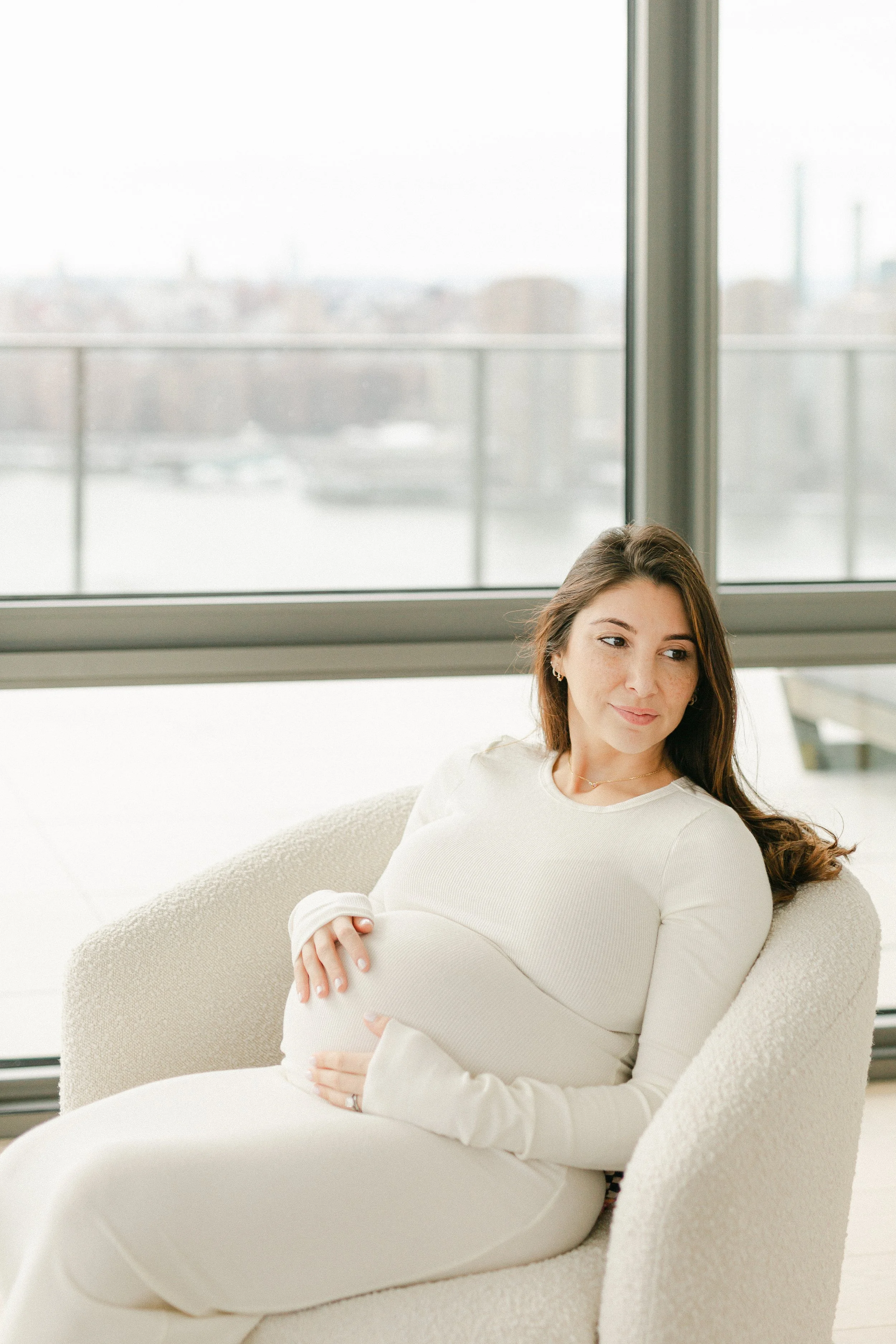 In-home lifestyle maternity portrait of a pregnant woman sitting on a cream-colored armchair near large windows with city view, gently holding her pregnant belly. Photographed by Maison Mancel in Brooklyn, New York.