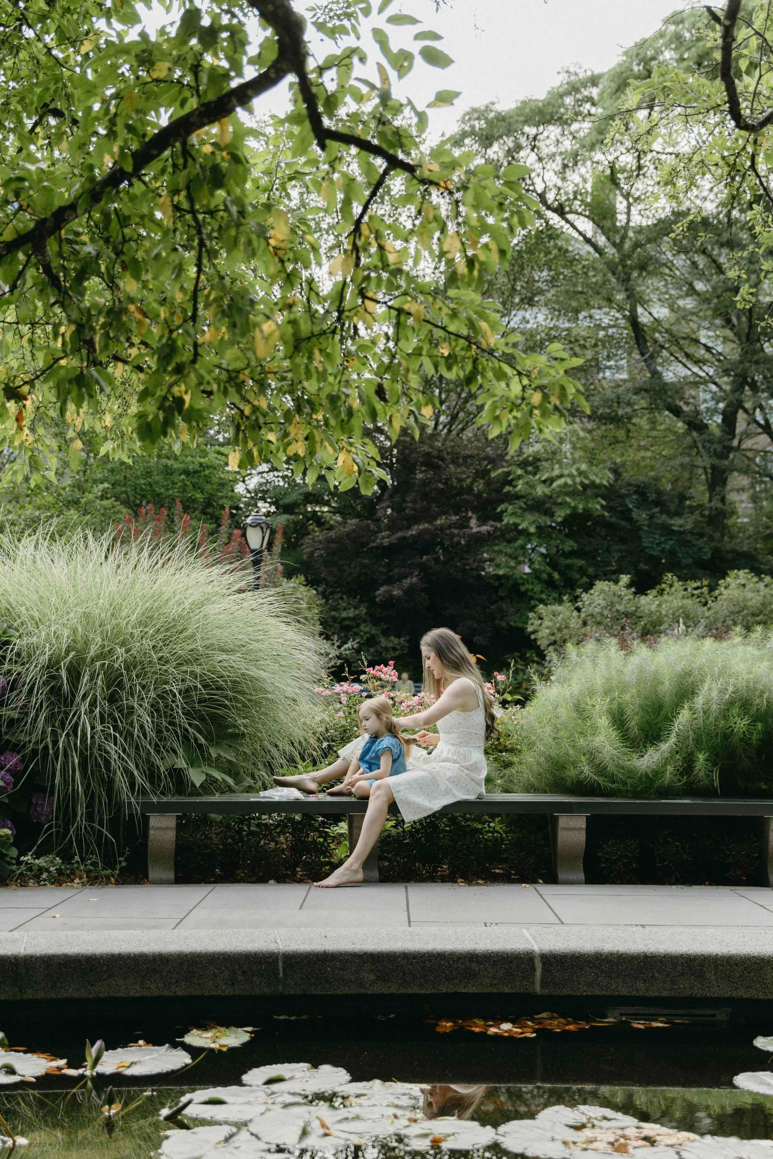 A mother sitting on a park bench with two children, surrounded by green plants and trees, near a small pond with lily pads in Central Park, by Maison Mancel.